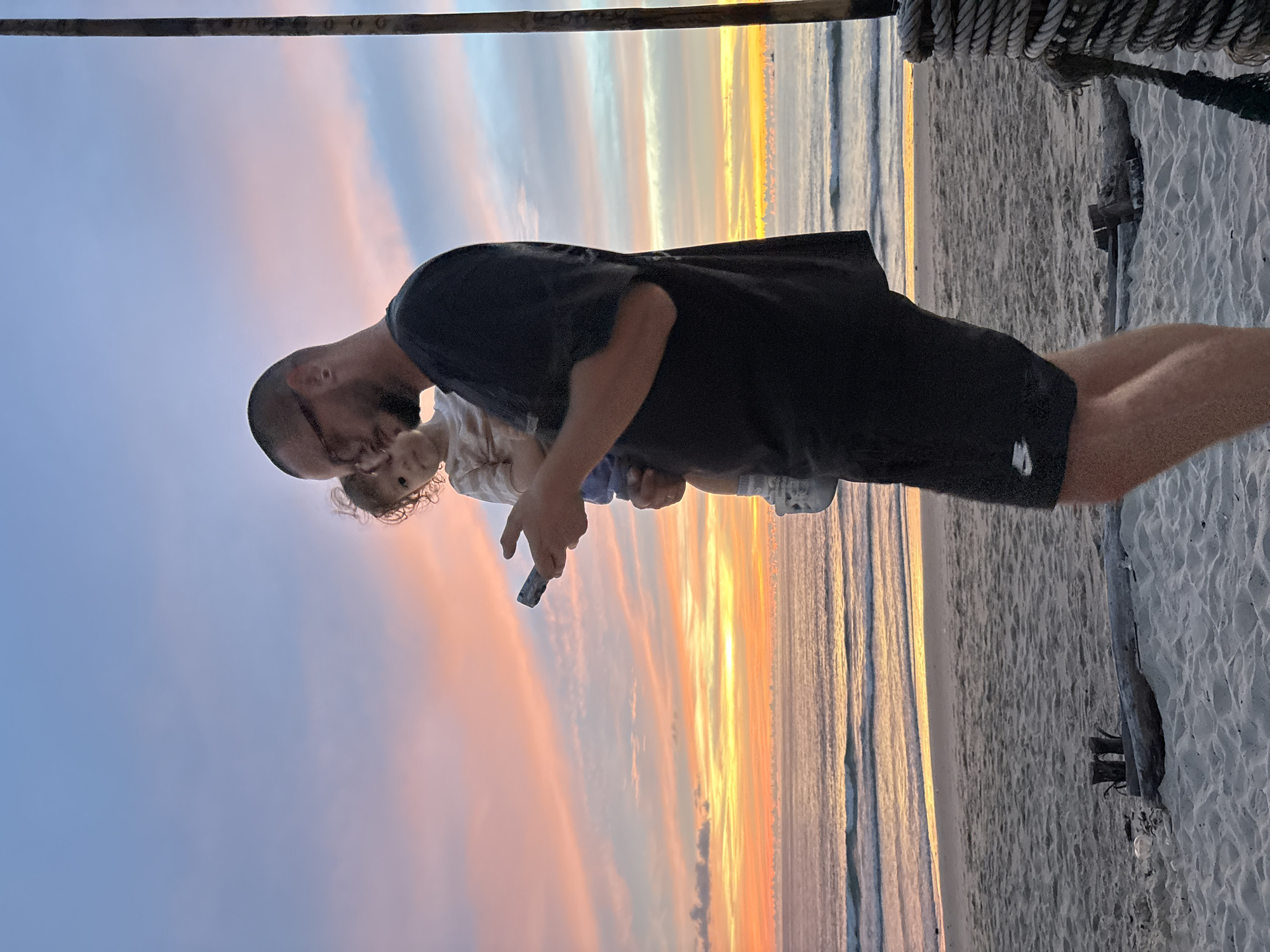 A man with glasses, wearing a dark t-shirt and dark shorts, holds a young child while looking at a mobile phone. The child, wearing a light-colored shirt, is nestled in the man's arms. They are standing on a sandy beach in Choeng Thale, Thailand, with the ocean and a colorful sunset in the background. The sky displays hues of orange, yellow, pink, and blue, with soft clouds. Waves are gently rolling onto the shore. In the foreground, a thick rope is coiled around a wooden post, and driftwood is scattered on the sand. The scene appears to be captured at dusk.