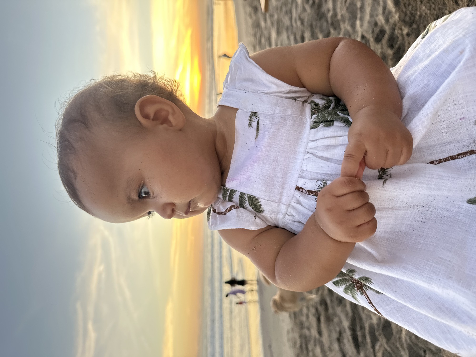 A baby in a white dress is playing on the beach at sunset in North Kuta, Indonesia. The baby's skin and dress are speckled with sand. There are people and a dog near the water.
