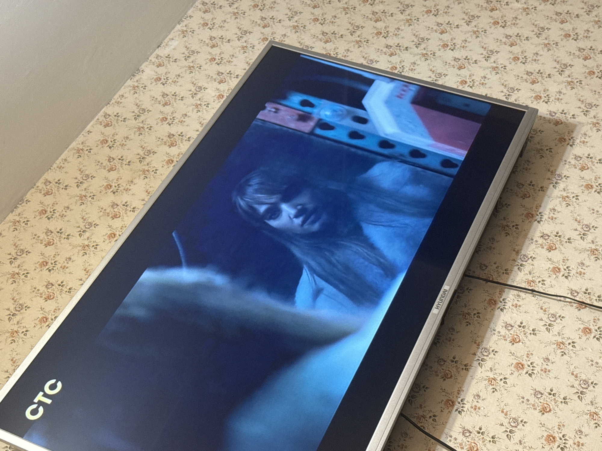 A television displaying a scene from a horror movie featuring a woman with long dark hair is resting on a surface covered with floral patterned wallpaper. The television brand is Hyundai. The channel logo CTC is visible in the bottom left corner of the screen. The location appears to be indoors. There is no information available to suggest that this photo was taken in Mytishchi, Russia.