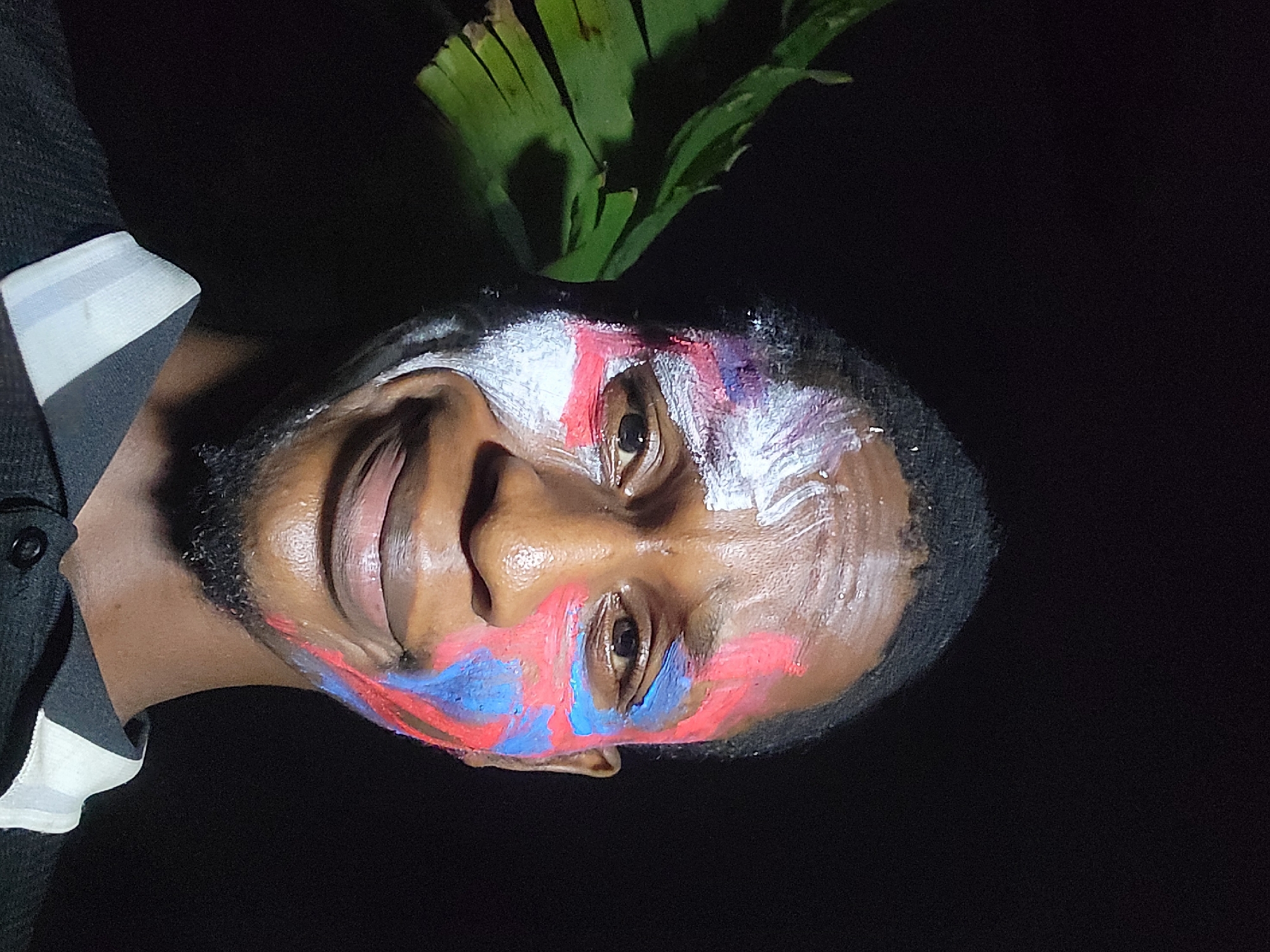 A medium shot depicts a person's face painted with blue, red, and white stripes. They are positioned against a dark background, with green foliage visible above their head. The person appears to be wearing a dark-colored shirt with a white stripe near the shoulder. This image was taken in Nkpor, Nigeria.