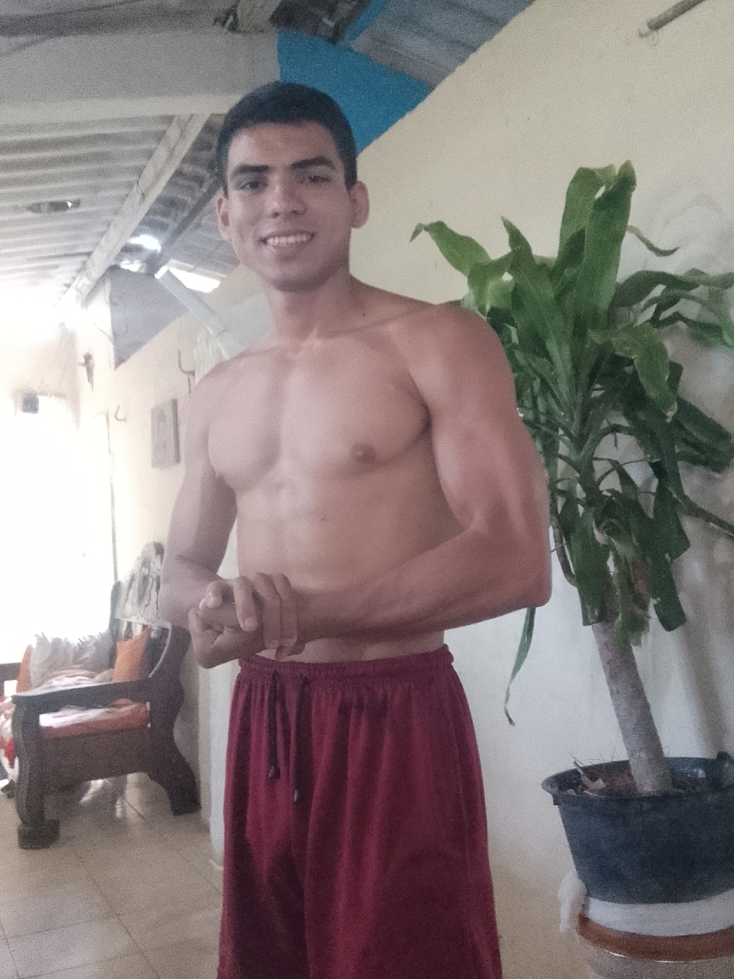 A shirtless, smiling, young man stands indoors wearing maroon drawstring pants. He is flexing his left arm and holding his hands together. Behind him to the right is a potted green plant. Behind him to the left is a window letting in bright light and a wooden chair with orange cushions. The interior space appears to have a concrete floor and a white wall. The location is likely a residential area within Juan Griego, Venezuela. The building has a weathered, slightly unkempt look.