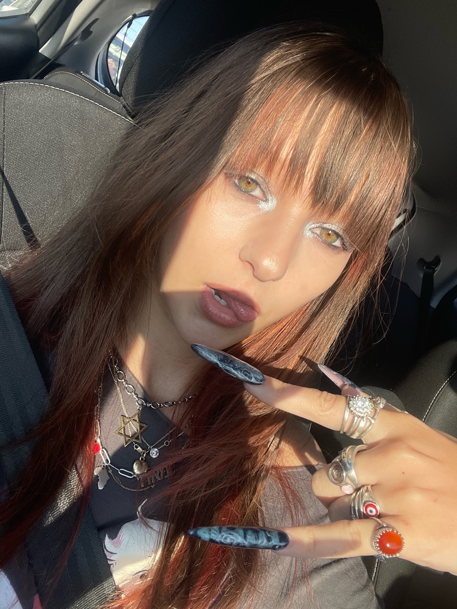 The image is a close-up, high-angle selfie of a young woman with long, reddish-brown hair and bangs, taken inside a car. She is wearing makeup, including silver eyeshadow and dark lipstick. She has long, pointed fingernails with a dark, swirling design and is adorned with multiple silver rings, including one with a large clear stone, another with an opal-like stone, and one with a red, evil eye design. She is also wearing several necklaces, including a star pendant and other charms. The woman is looking at the camera with her tongue slightly out and is making a rock-and-roll hand gesture. The lighting suggests it is daytime, with sunlight casting shadows on her face and hair. The background shows the interior of a car, including a dark seat and a glimpse of the window with sunlight streaming through. There is no indication of the city of Raanana, Israel in the image itself.