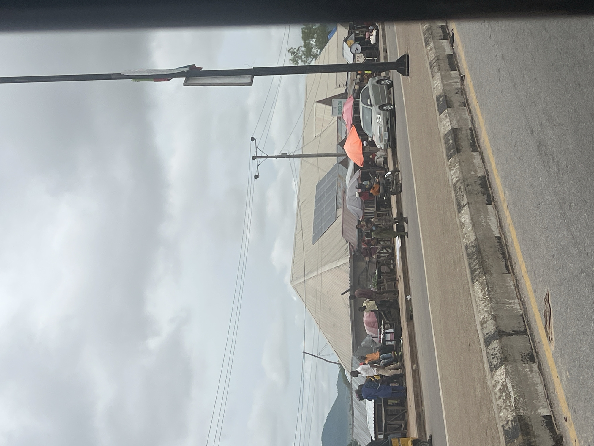 The image depicts a busy street scene in Jalingo, Nigeria, on an overcast day. A roadside market is set up with various stalls covered by umbrellas. People are seen interacting with vendors and browsing goods. Several vehicles, including a silver sedan and a motorcycle, are present on the road. The background shows a range of buildings, some with corrugated metal roofs, and a lush green hillside in the distance. A prominent utility pole with wires is visible on the right side of the frame, along with a lamppost. The street is paved with asphalt and has a concrete curb separating it from the market area.
