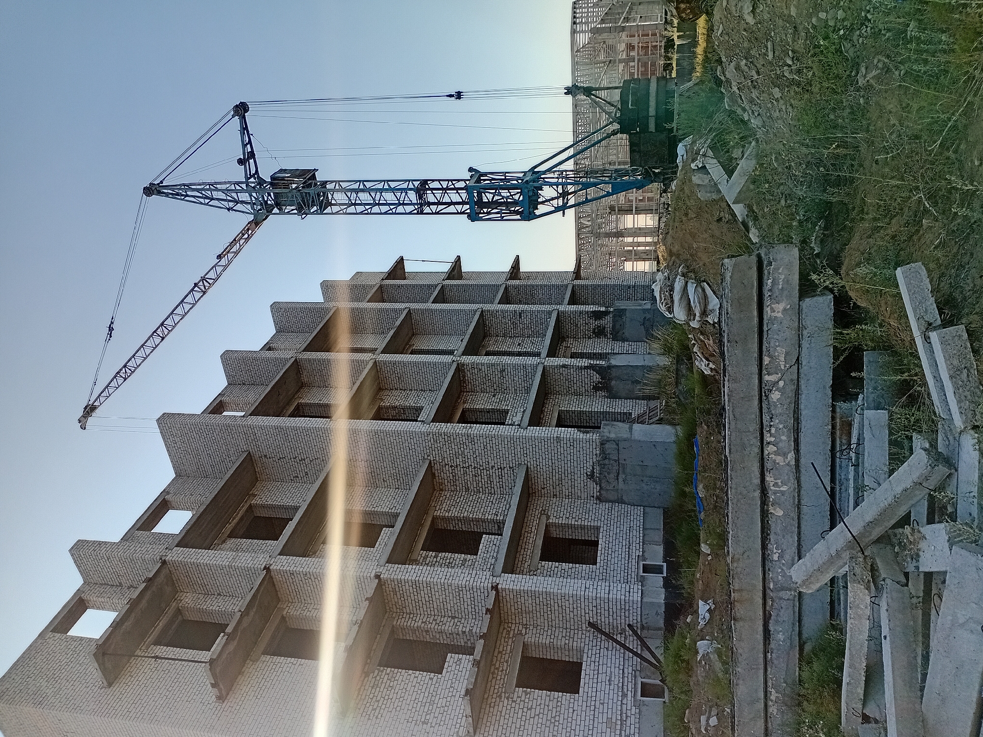 The image captures a construction site in the city of Semey, Kazakhstan. In the foreground, a pile of concrete slabs and debris is visible, with overgrown grass and dirt surrounding them. A multi-story building, constructed with white bricks and concrete balconies, is in the process of being built, with its windows and doorways open and unfinished. A large blue construction crane stands tall beside the building, extending its boom over the site. In the background, the skeletal framework of another structure is under construction, suggesting a larger development project. The sky is clear and pale blue, indicating daytime. There are no people visible in the image.