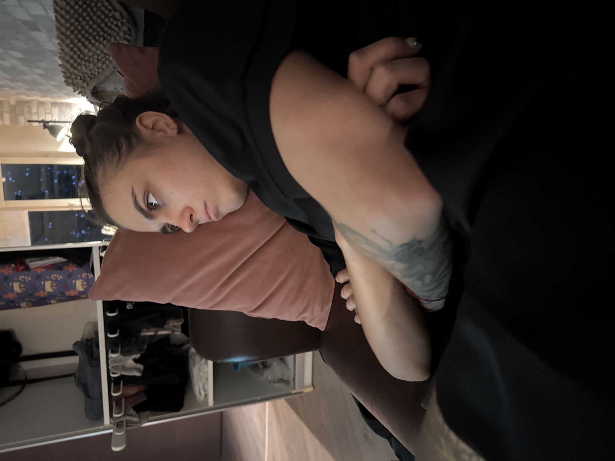 The image shows a young woman with her hair pulled back in a bun, looking to her left with a pensive expression. She is wearing a black shirt and appears to be lying down, possibly on a couch or bed, with her arms crossed. A pink pillow is behind her head, and a dark blanket or comforter is draped over her. Tattoos are visible on her arm. In the background, there is a shelf with clothes, a lamp, and a window with lights visible outside. The scene suggests an indoor setting, likely a residential space. The image does not provide any information about the location being Moscow, Russia.