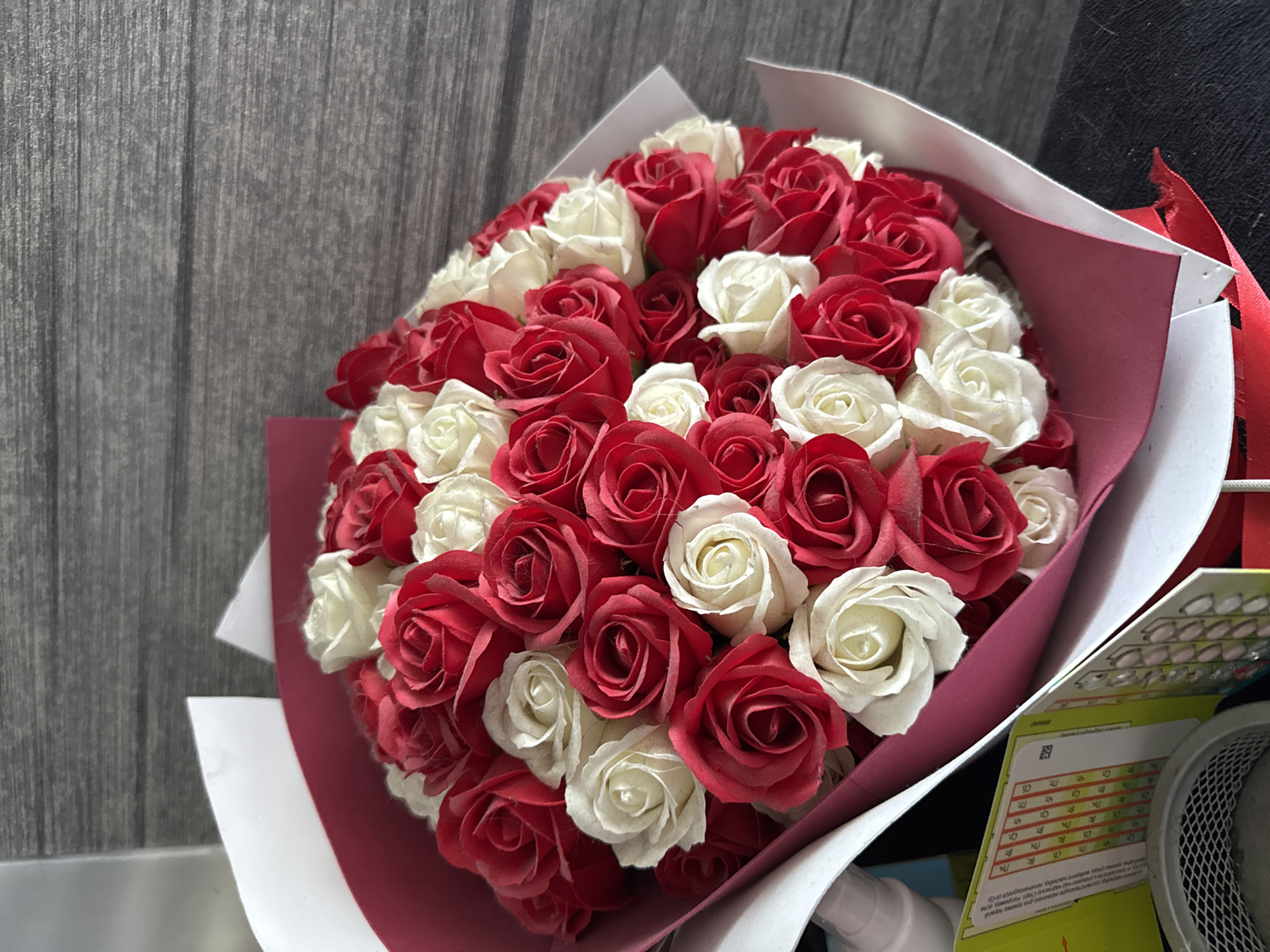 This media file features a close-up view of a bouquet of red and white roses, artistically arranged. The roses, appearing to be artificial, are tightly packed, creating a full and vibrant display. The bouquet is partially wrapped in white and deep pink paper, with a red ribbon peeking out from the side. Behind the bouquet, a textured grey background, possibly a wooden wall or paneling, is visible. To the right of the bouquet, a portion of a desk or table is shown, with a metal mesh container and what appears to be packaging for medication or vitamins. The visible text on the packaging is in a foreign language and includes numbers and symbols, such as "18," "2," "D," "N," "BT," "Чт.," "10," "19," "BC," and "32." The lighting suggests an indoor setting, possibly a room or office, with no specific time of day or weather conditions indicated. The overall impression is of a carefully presented gift, likely intended for a special occasion.