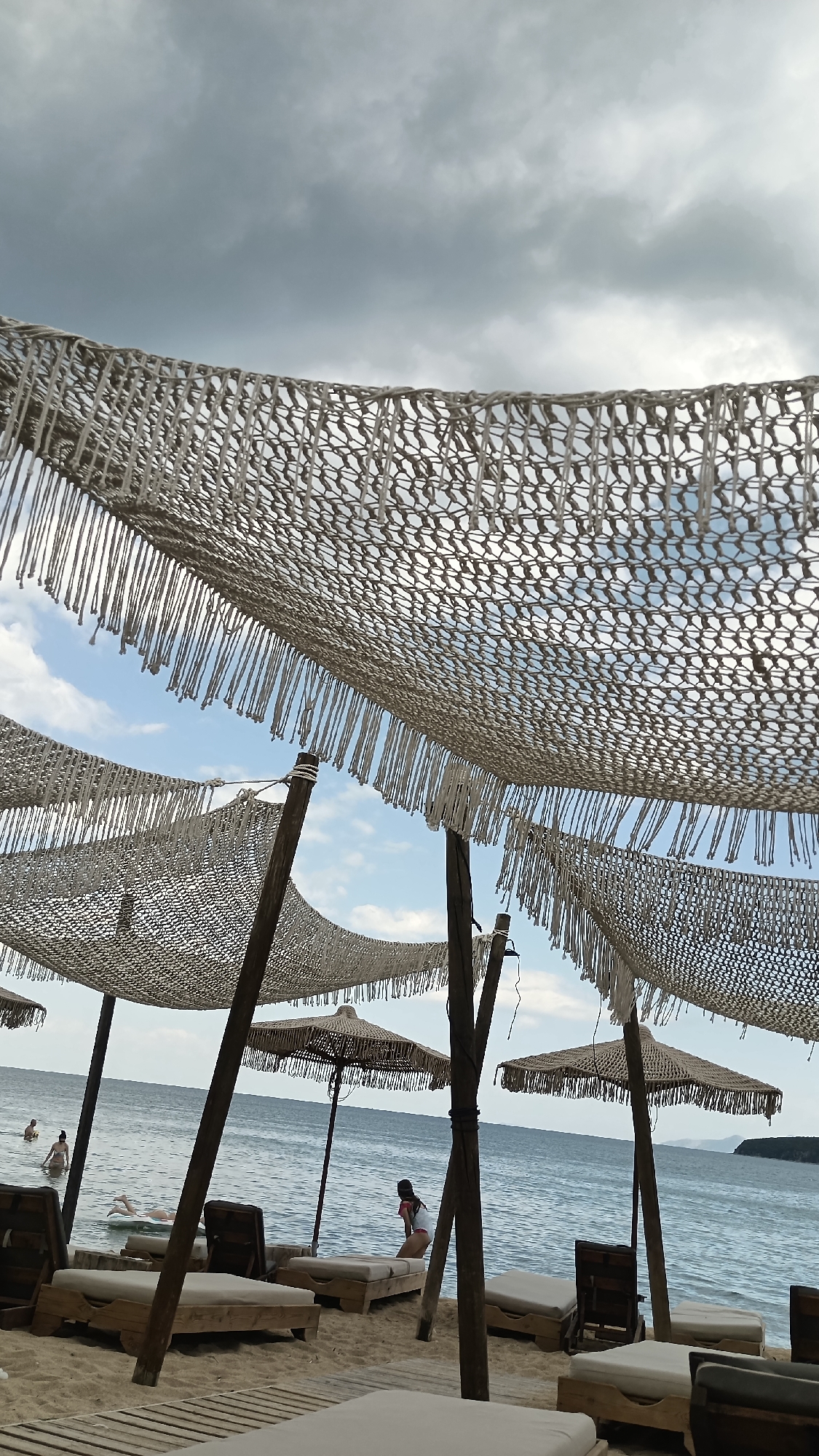 A beach resort in Nea Peramos, Greece, features several rustic, open-air cabanas with woven canopies and fringed edges, supported by wooden poles. Underneath these canopies are rows of lounge chairs, many of which are unoccupied. The beach itself is sandy, with wooden platforms laid out in some areas. The sea is visible in the background, with a few people swimming or wading in the water. One person is floating on a white inflatable raft, while another is sitting on a lounge chair near the water's edge. The sky is overcast with dramatic clouds.