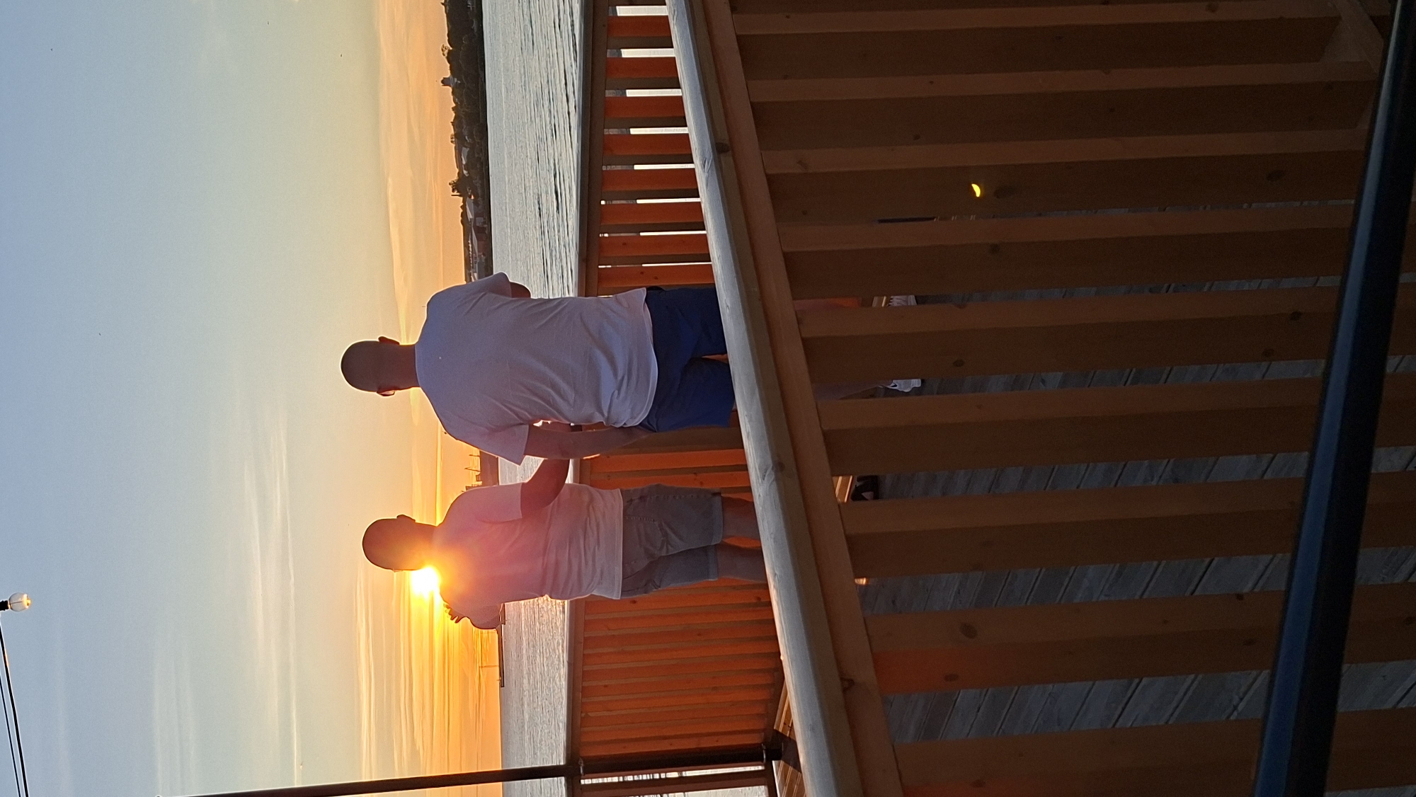 Two men stand on a wooden deck, facing a sunset over a body of water. The man on the left wears a light-colored t-shirt and grey shorts. The man on the right wears a white t-shirt and blue shorts. The sky is a gradient of orange and blue, with a bright sun near the horizon. The deck has a wooden railing in the foreground. The background shows a distant shoreline with buildings and trees. The scene is located in Tallinn, Estonia, near the Seaplane Harbour Museum.
