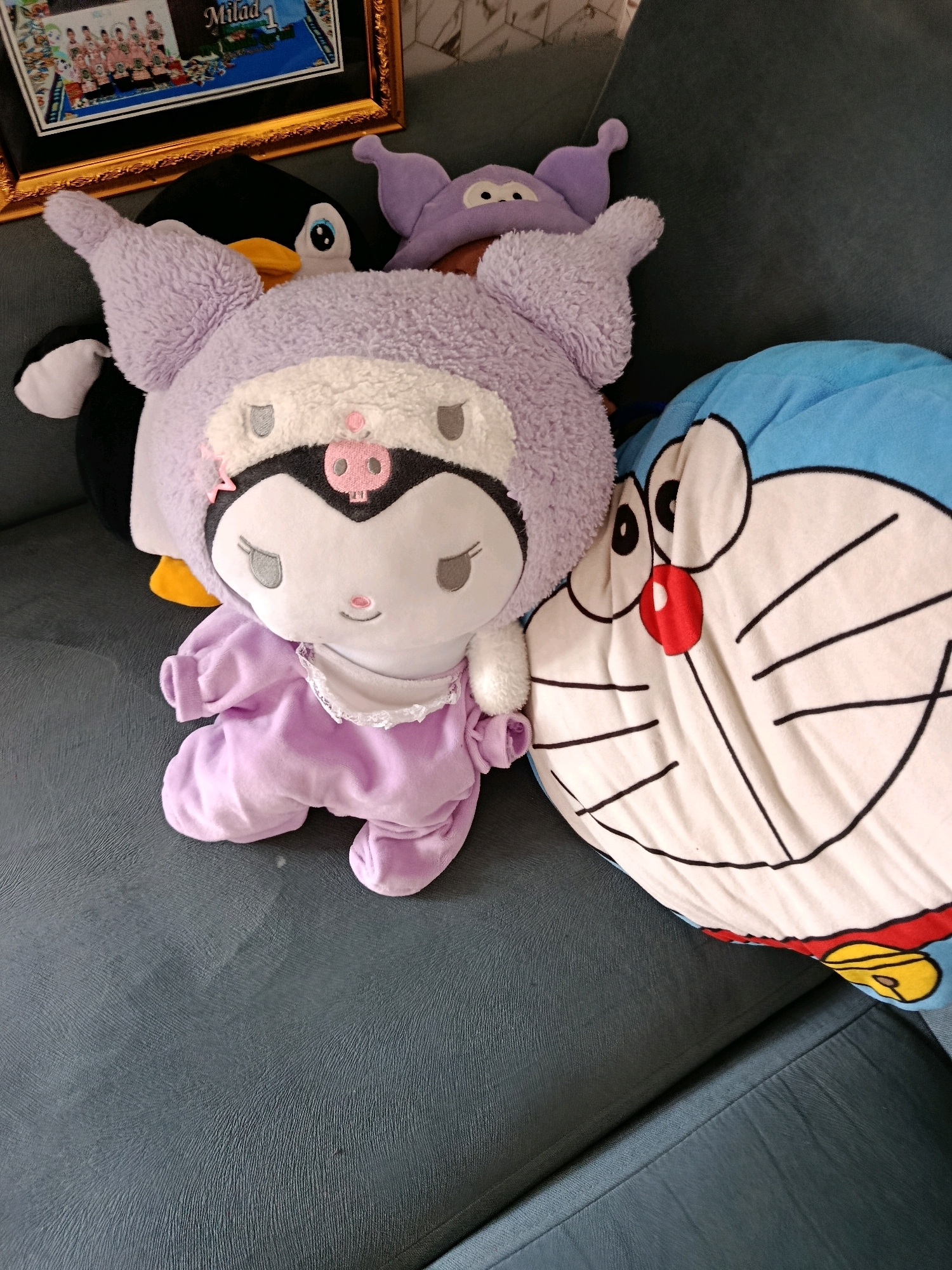 In the city of Karang Rejo, Indonesia, a dark grey couch is occupied by several plush toys. A large blue plush toy with the likeness of Doraemon is positioned on the right side of the couch. To the left of Doraemon, a lavender Kuromi plush toy is seated, wearing a lavender outfit. Behind Kuromi, a black penguin plush toy with yellow accents is visible. Partially obscured behind the penguin is a small, lavender, bat-like plush toy with large eyes. In the upper left corner of the image, a framed picture rests on a surface above the couch. The frame is gold and ornate, containing a photograph of a group of people. The lighting in the image is even, suggesting an indoor setting.