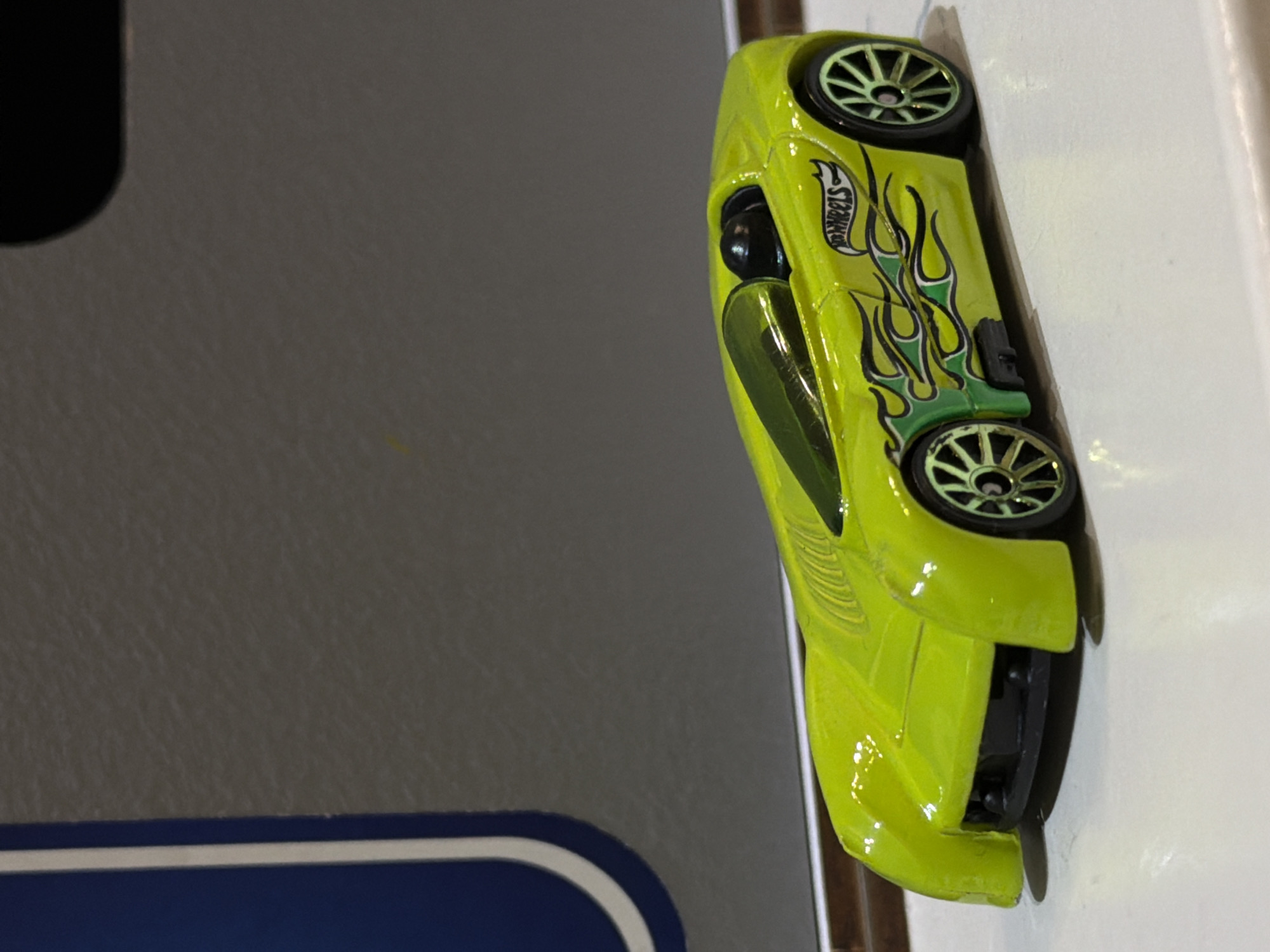 The image captures a close-up, vertically oriented view of a bright lime green Hot Wheels toy car, positioned on its side. The car's body features distinct black and green flame decals, with the 'HOT WHEELS' logo visible amidst the design on its upper side. It is equipped with black wheels that have prominent green spokes. The car has a glossy finish, reflecting some light. The background on the left is a textured matte grey surface, while the right side is a plain white surface. A dark blue band with a thin white border is partially visible at the bottom left. There is no discernible activity; the car is stationary, suggesting an indoor setting. This media file was captured in Sofiivska Borshchahivka, Ukraine.