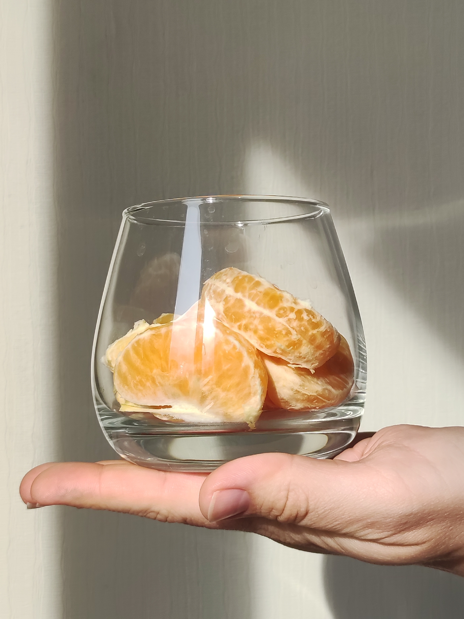 A hand holds a clear glass filled with orange segments against a light-colored, textured wall. The image focuses on the hand and the glass. The orange segments are visible inside the glass. The lighting is bright, creating highlights and shadows. The environment appears to be indoors.