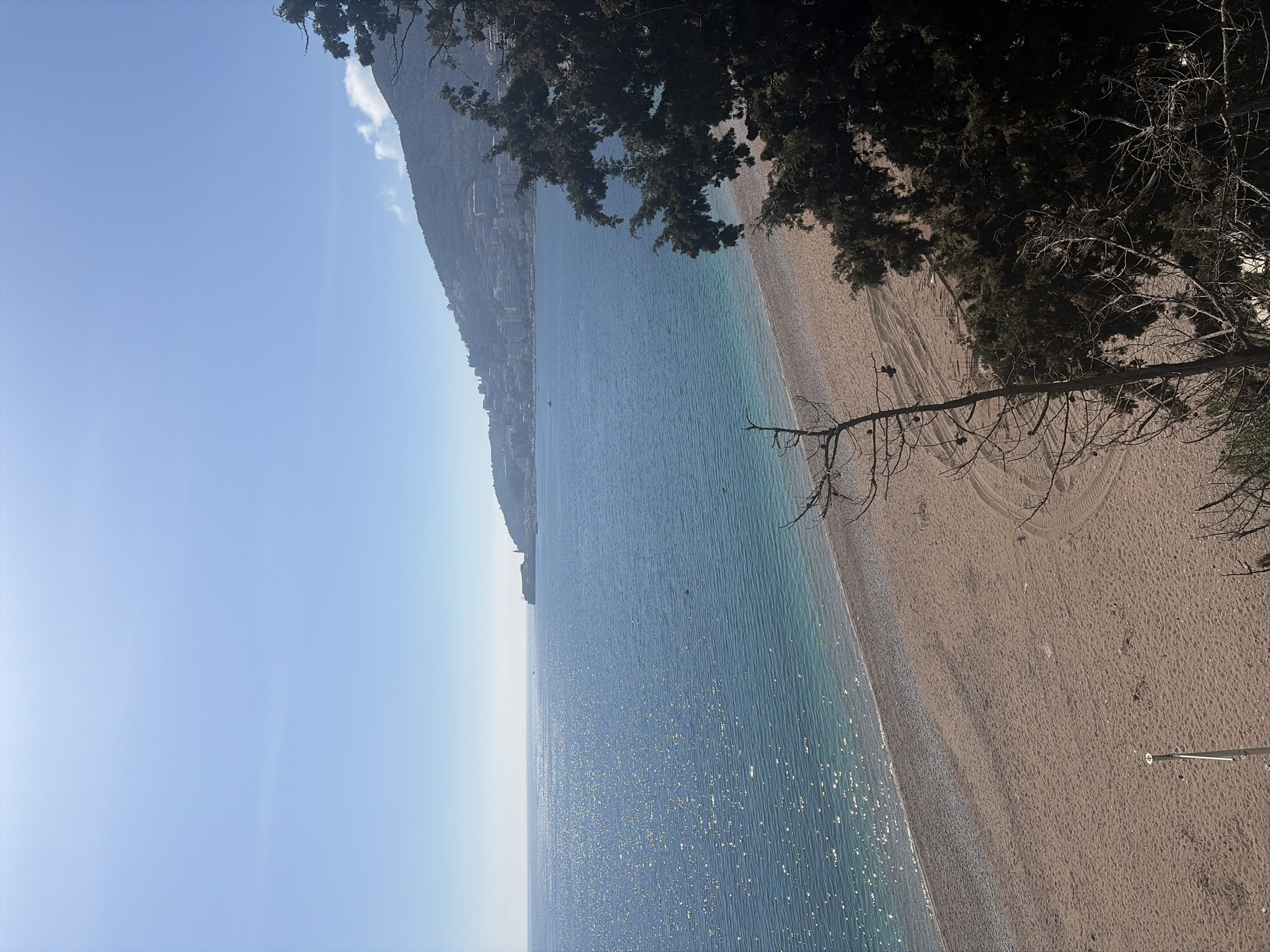 The image shows a beach in an unknown city. The sea is a sparkling blue with sunlight reflecting off the surface. A long beach stretches along the edge of the water. There are trees on the far right of the image. A mountain range extends into the horizon on the left.