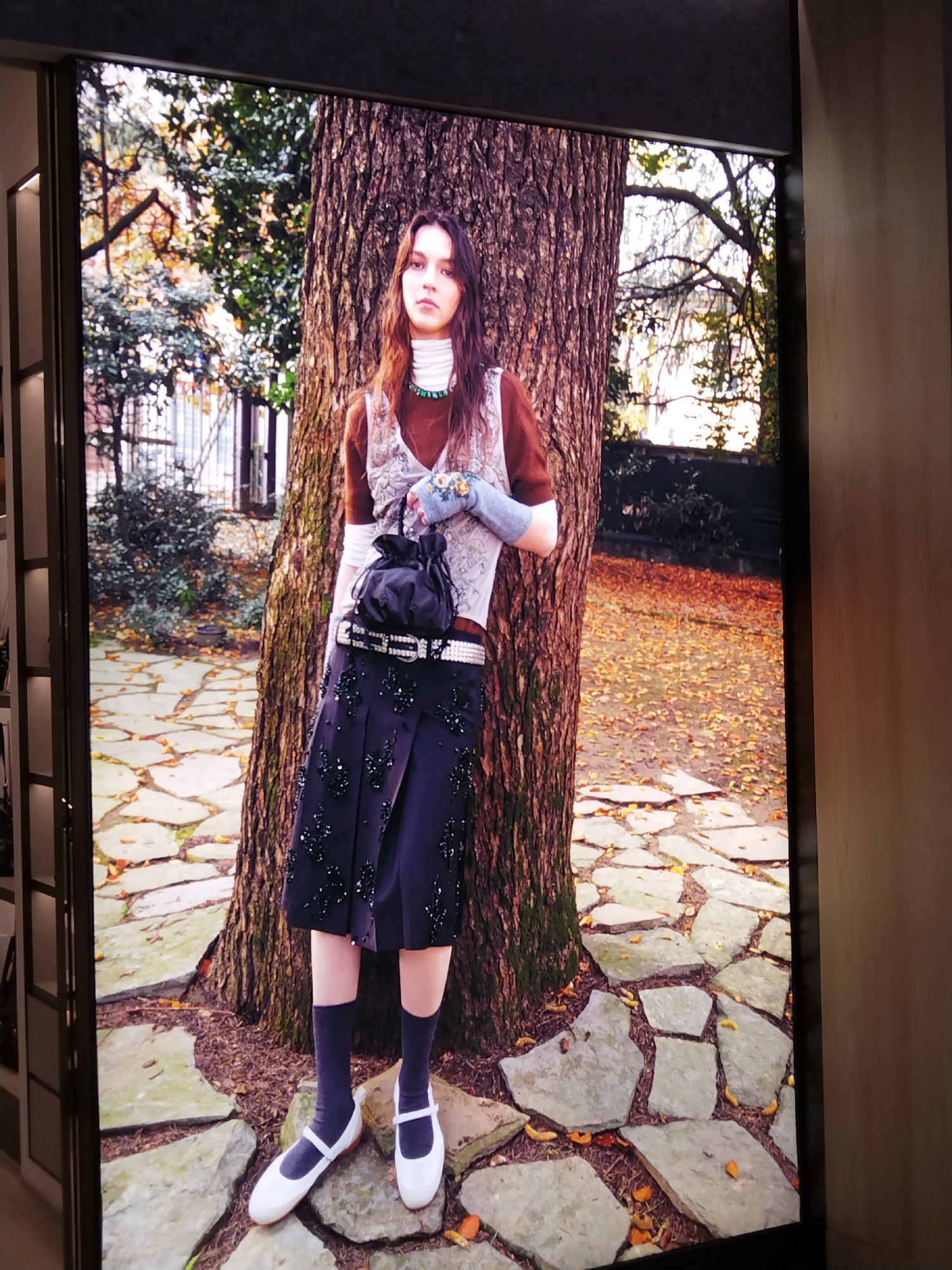 The image shows a digital display featuring a fashion advertisement. A young woman is depicted standing next to a large tree in an outdoor setting with fallen leaves on the ground and stone pathways. She is wearing a brown long-sleeved shirt under a patterned vest, a dark skirt embellished with beads, white shoes, and dark socks. She is also holding a black drawstring bag and wearing a blue bracelet on her left arm. The background shows trees and foliage, suggesting an autumnal environment. The display itself is framed and appears to be in a dimly lit interior space, possibly a store or exhibition hall. There is no indication that this scene is located in Patong, Thailand.