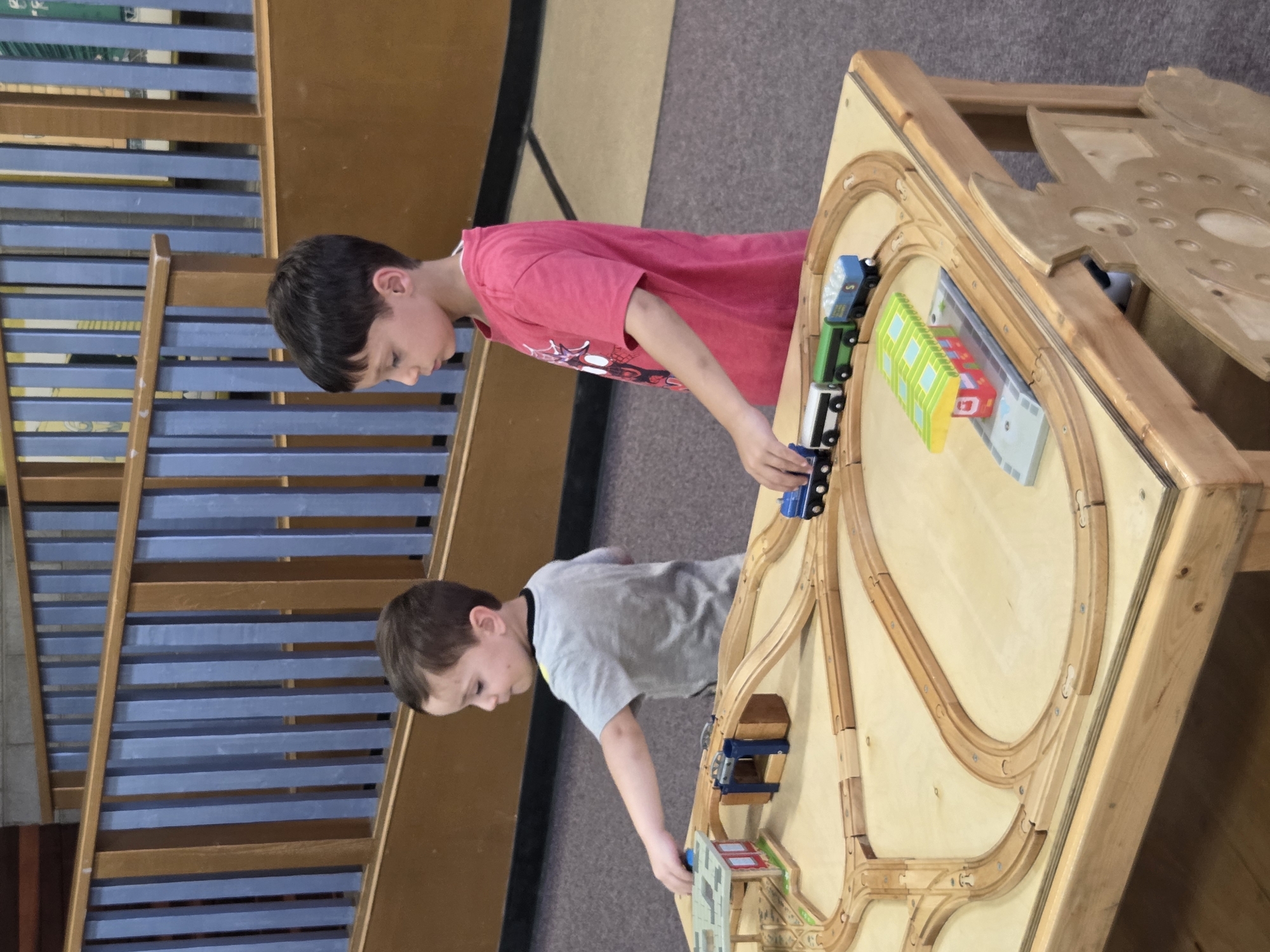 Two young boys are playing with a wooden train set on a table in what appears to be a community center or children's museum. The boy on the left, wearing a grey t-shirt, is reaching out towards the train track. The boy on the right, wearing a pink t-shirt with a graphic on it, is looking down at the train set. The train set includes wooden tracks, bridges, a station, and several toy trains. The table is made of wood, and the surrounding environment has wooden railings and carpeted floor. There is no indication of the specific location within Los Angeles, United States of America.