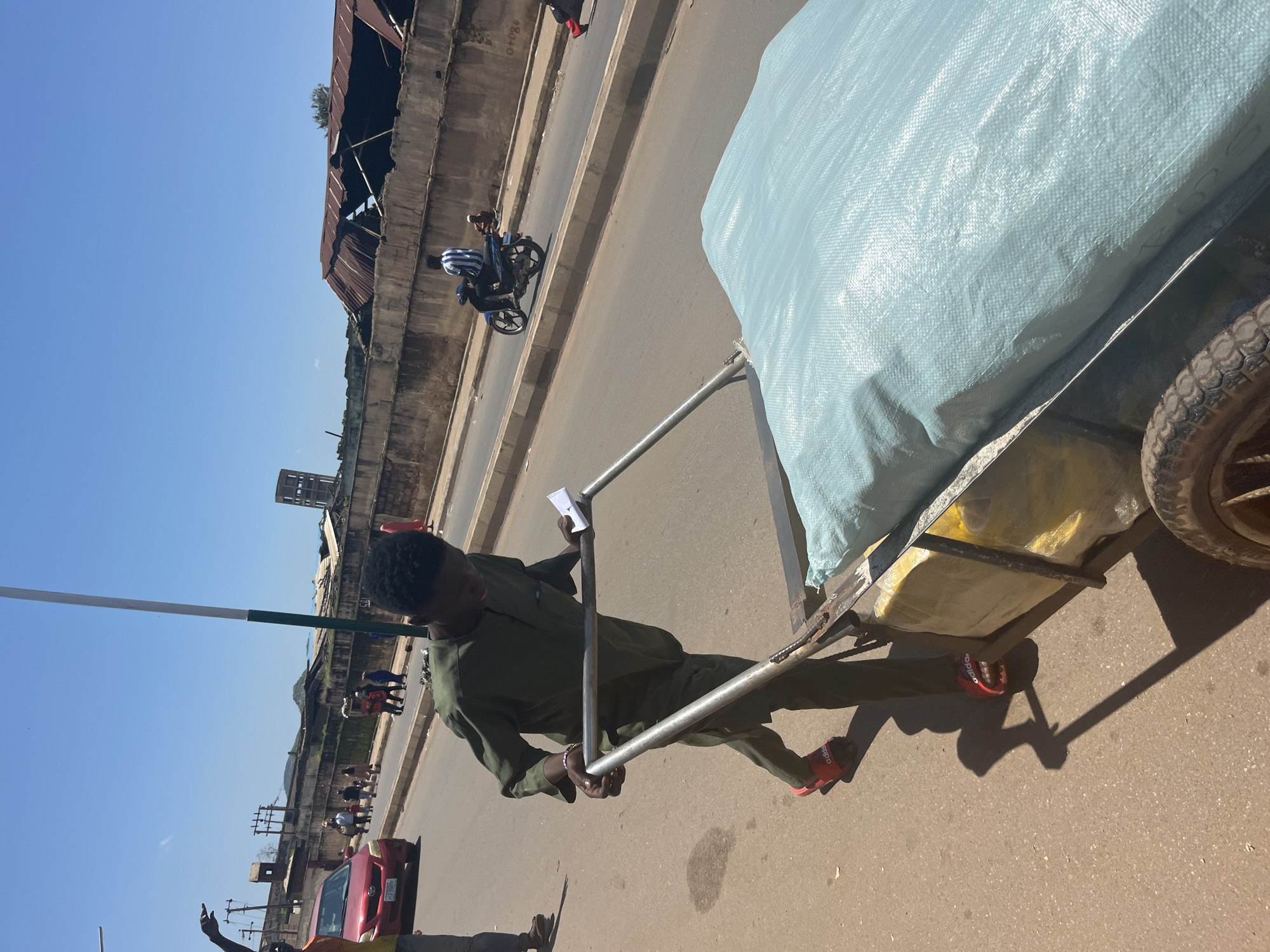 In Jos, Nigeria, a young man dressed in a green traditional tunic and red Adidas slide sandals is pushing a handcart laden with a large, light blue-covered load. The handcart appears to be made of metal and has two wheels. The man is looking down at a piece of paper he is holding in his right hand, with his left hand gripping the cart's handle. The scene unfolds on a paved street, with a concrete wall bordering the right side of the road, showing signs of disrepair. Beyond the wall, structures with corrugated iron roofs are visible. In the background, several people are walking along the sidewalk, and a red car is parked on the side of the road. A motorcycle with two people on it is also present on the road. The sky is clear and blue, indicating a sunny day. The overall environment suggests a bustling urban setting.