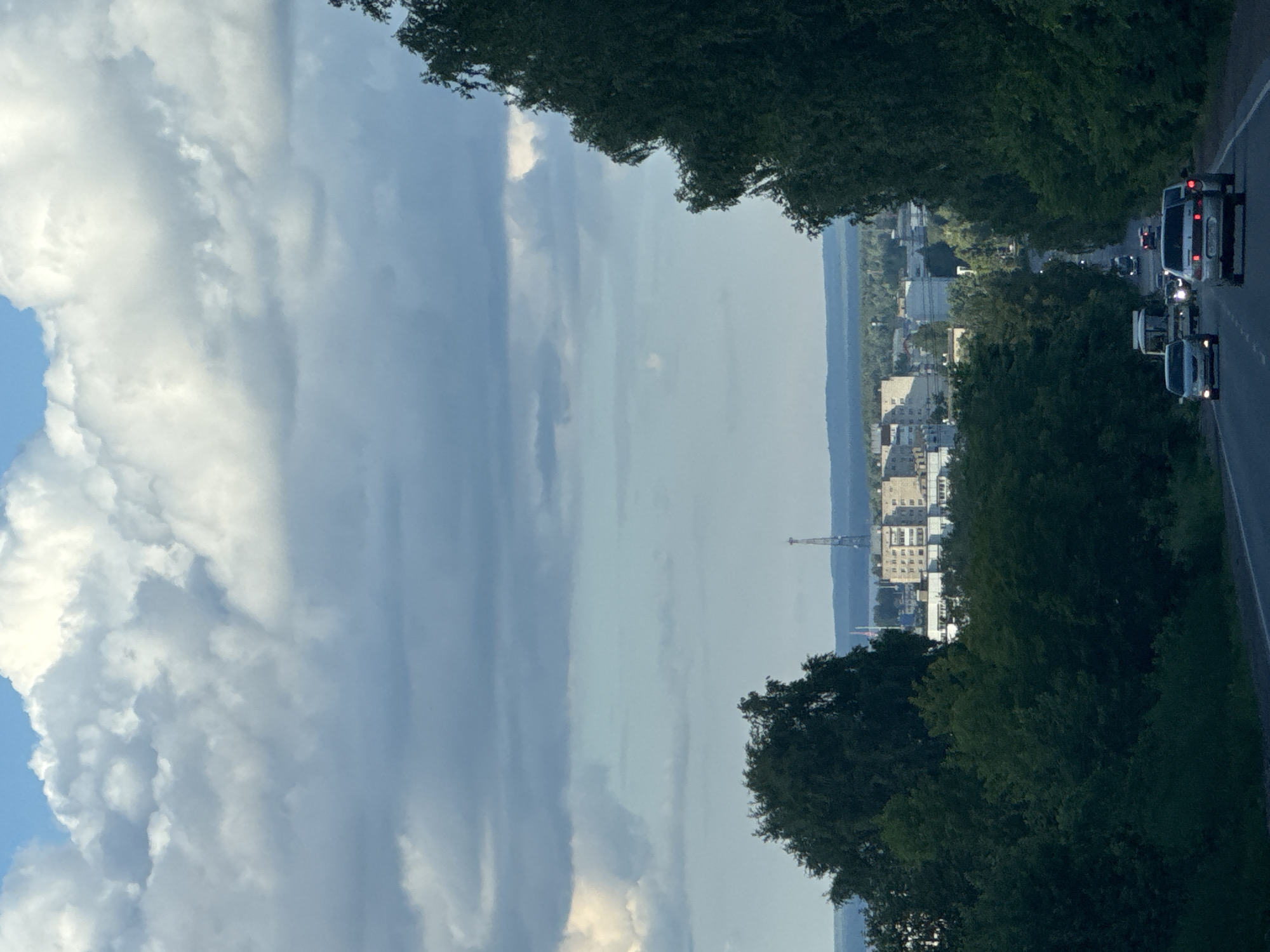 The image captures a scenic view of a road leading towards the city of Chaikovsky, Russia. The sky is filled with dramatic, voluminous white and gray clouds, with patches of blue sky visible. A tall, lattice-like transmission tower stands prominently in the distance, above a cluster of buildings that form part of the city's skyline. The road is flanked by dense, green trees on both sides, creating a tunnel-like effect. Several cars are visible on the road, moving away from the viewer, including a white van, a light-colored sedan, and another sedan with its brake lights illuminated. The overall impression is one of a journey through a lush, natural landscape towards an urban center under a dynamic sky.