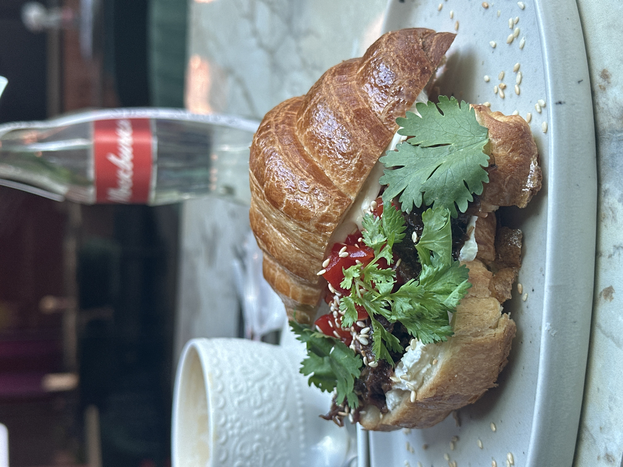 A croissant sandwich is positioned on a light grey plate with scattered sesame seeds. The sandwich appears to be filled with cream cheese, a dark meat product, diced red bell peppers, and sprigs of cilantro. A bottle of water with a red label and a white ceramic cup with a detailed pattern are partially visible in the background. The location is Novosibirsk, Russia.