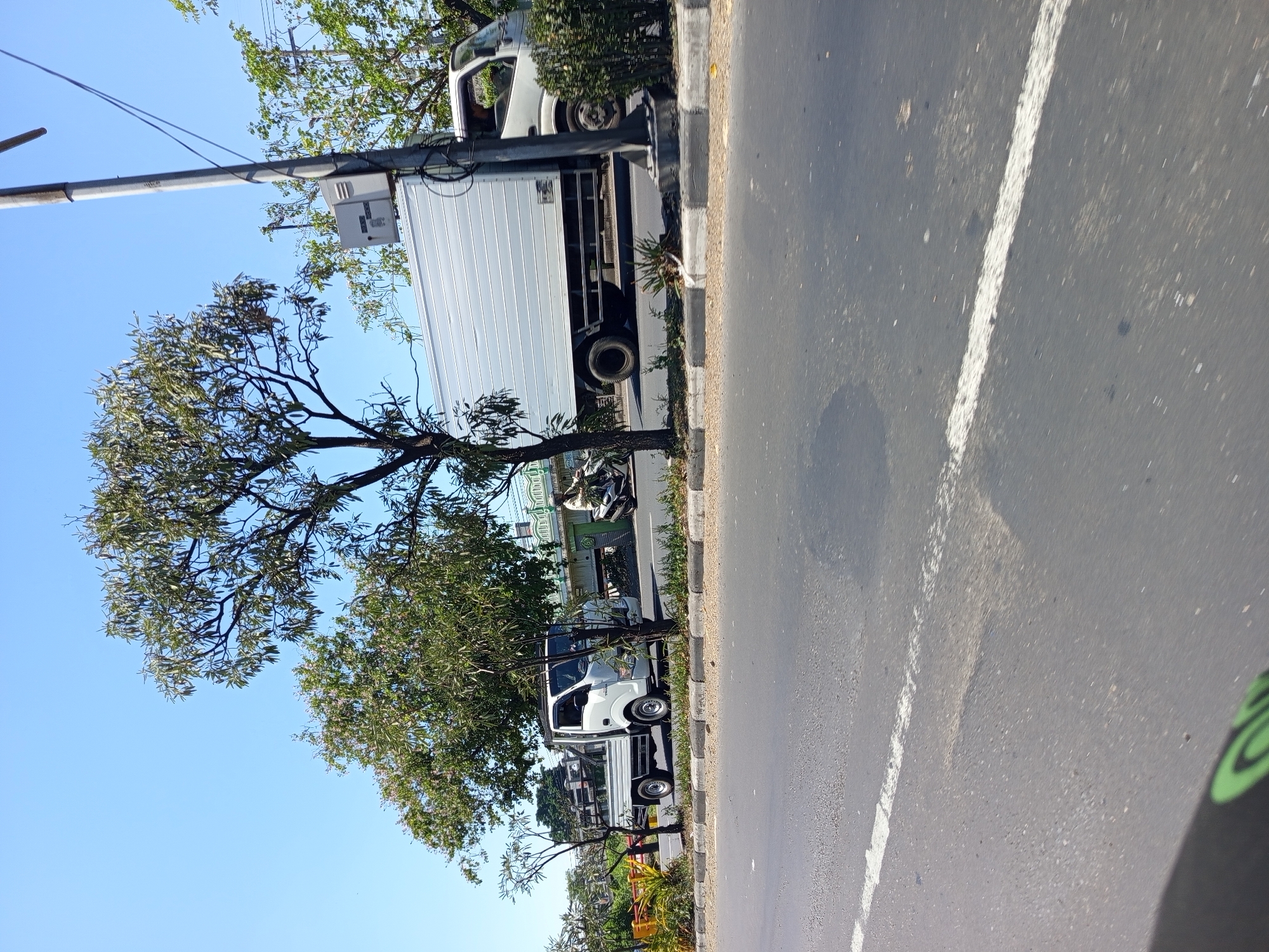 In Sawotratap, Indonesia, under a clear blue sky, a white pickup truck with a silver cargo box is traveling on a paved road. To the left of the pickup truck is a smaller white truck with an open bed, also on the road. A motorcycle with a rider is visible between the two trucks. The roadside is lined with trees and a sidewalk. In the foreground, the asphalt road shows a white lane marking.