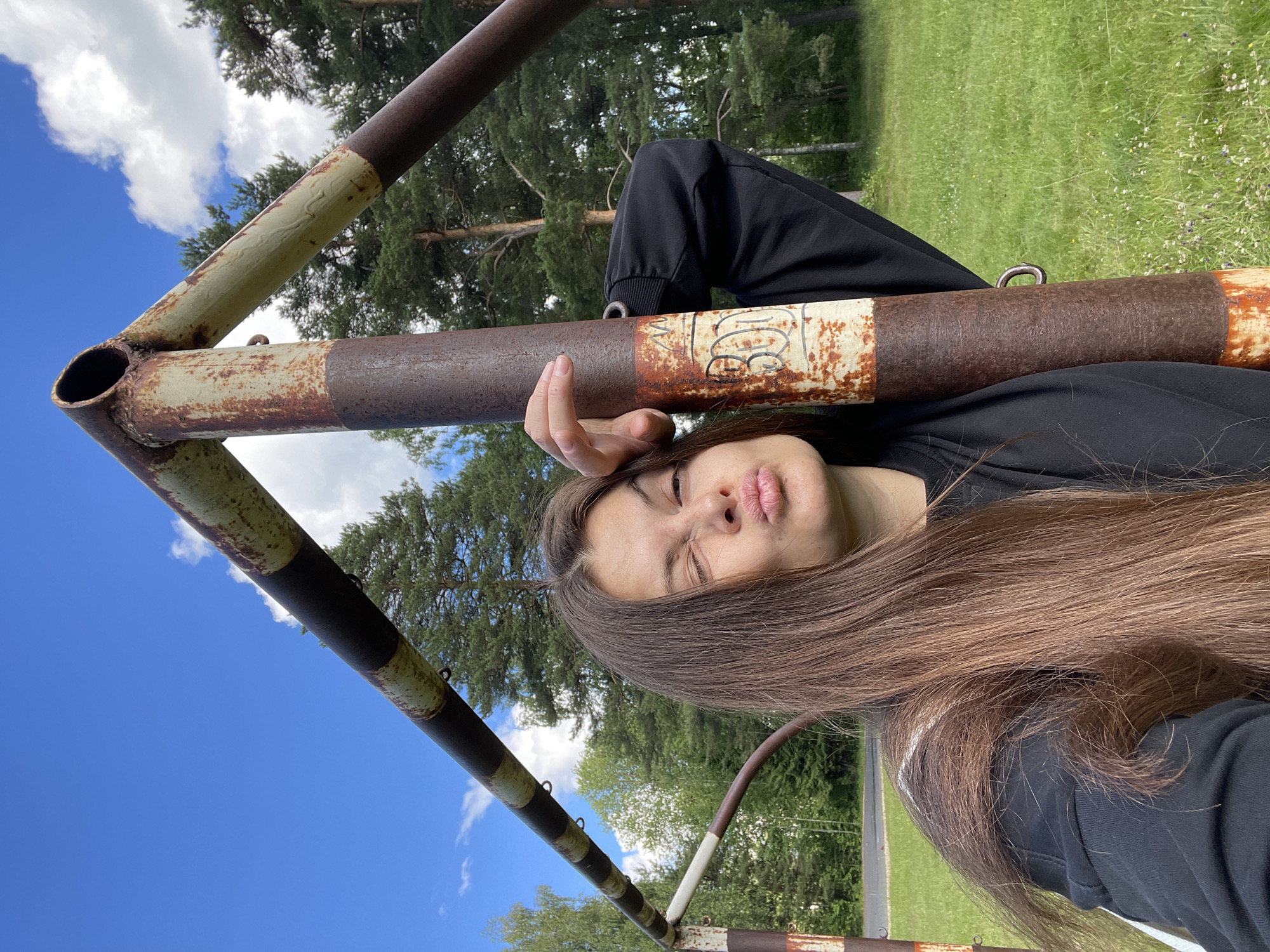 A young woman is lying down outdoors, resting her head on a rusty metal bar. She is wearing a black hoodie. Her eyes are closed, and her lips are puckered, as if she is about to blow a kiss or make a kissy face. Her long, brown hair is spread out around her head.
The setting appears to be a grassy field with trees in the background, suggesting a park or a rural area. The sky is blue with some white clouds, indicating it is daytime and likely pleasant weather. The metal bar she is lying on looks like part of an old piece of playground equipment or sports equipment, such as a goalpost or a swing set. It is weathered and shows signs of rust.
There is some text visible on the metal bar, but it is difficult to read clearly. It appears to be handwritten in black marker.
The overall mood of the image is relaxed and playful, with the woman's expression and pose conveying a sense of lightheartedness. The orientation of the image is rotated, making it appear sideways.