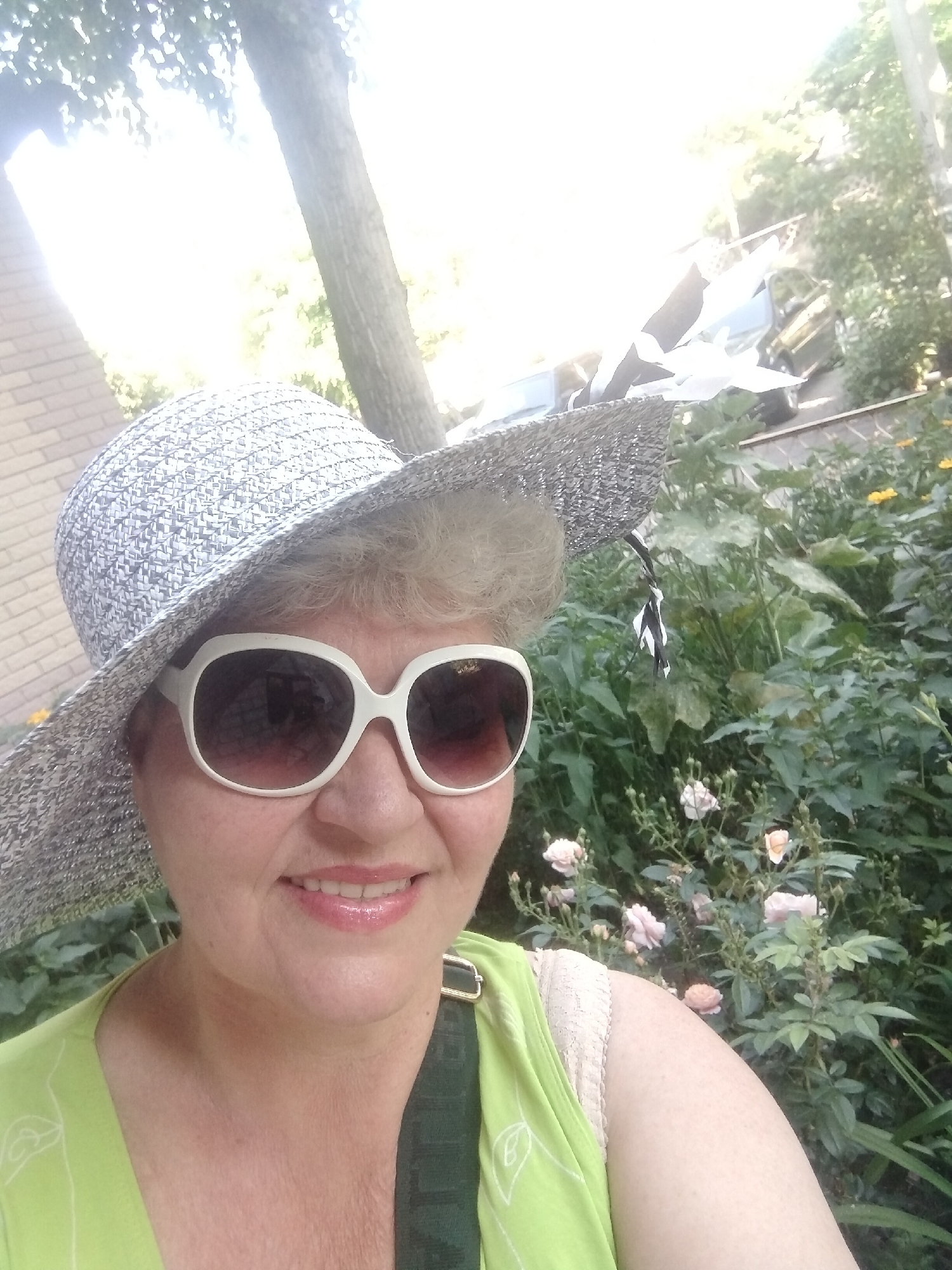 A selfie taken outdoors shows a woman wearing a wide-brimmed hat and large white-rimmed sunglasses. She is smiling and her lips are painted with pink lipstick. She is wearing a lime green top with a dark strap visible across her shoulder. The background consists of lush green foliage and rose bushes with pink flowers. A brick building is visible to the left and what appears to be a car is in the background to the right. The lighting suggests it is daytime, possibly with bright sunlight filtering through trees. The presence of rose bushes and a garden setting suggest this could be a residential backyard or a park area within Horishni Plavni, Ukraine.