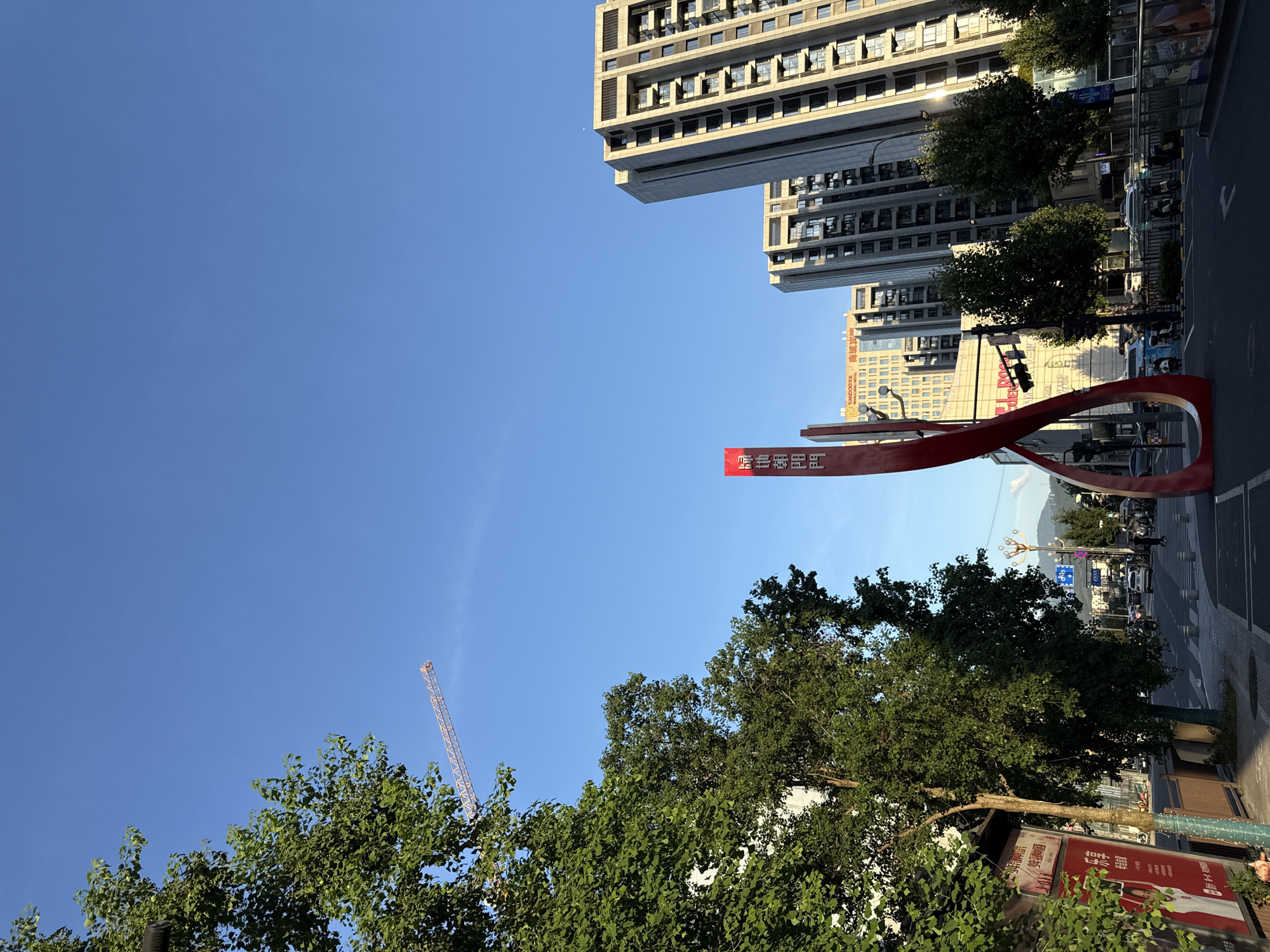 The image depicts a street scene in Yiwu, China, under a clear blue sky. In the foreground, a distinctive red abstract sculpture, possibly an art installation or landmark, dominates the center of an intersection. To the left, lush green trees overhang the sidewalk, partially obscuring a building with a red advertising board featuring Chinese text and an image of a woman. Further down the street, a traffic light hangs over the intersection, controlling the flow of vehicles.
On the right side of the image, several tall, modern residential buildings with multiple balconies rise into the sky. Between these buildings, more trees can be seen, as well as a section of another building with a yellow facade and Chinese characters. In the distance, a construction crane is visible against the blue sky, indicating ongoing development in the city. The street is paved, with visible lane markings and pedestrian crossings. Several cars and motorcycles are present on the road, with a few pedestrians walking on the sidewalks. The overall atmosphere suggests a typical urban environment in Yiwu during daylight hours, with a blend of modern architecture, greenery, and everyday city life.