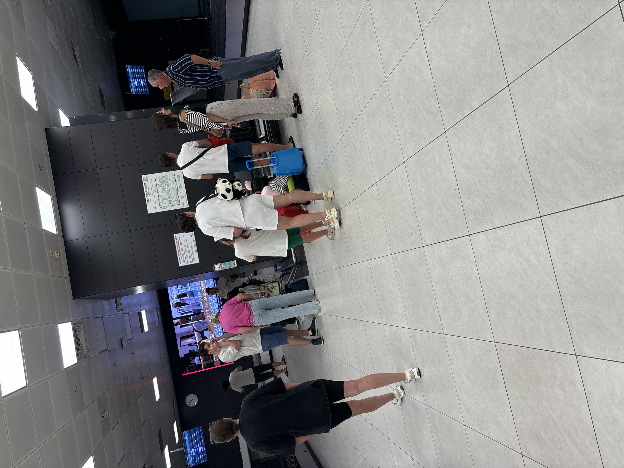 In the city of Malye Kabany, Russia, an airport terminal is depicted with a group of people standing and interacting. The scene is captured from a slightly elevated perspective, looking down and across a tiled floor.
In the foreground, a woman in a black t-shirt and black shorts walks away from the camera, facing left. She has sunglasses on her head and is wearing white sneakers.
To the left, a man with dark hair, wearing a grey t-shirt and blue shorts, is speaking on his phone. Next to him, a woman in a bright pink t-shirt and light blue jeans stands facing away from the camera. Behind them, a man in a white t-shirt and dark shorts also stands.
In the mid-ground, a group of people are gathered. A woman in a white t-shirt and white shorts stands facing right, holding a stuffed panda. Beside her, a young girl in a white t-shirt and green shorts is being spoken to by a woman in a black and white striped top and light-colored pants. A blue suitcase is on the floor near them. Further to the right, a man in a striped blue and white shirt and grey pants is talking to another man in a white t-shirt and dark shorts. Several bags are visible near this group.
The environment appears to be an indoor space with a high ceiling featuring multiple fluorescent lights. A large screen displaying flight information or advertisements is visible on the left wall. A clock is also mounted on the wall. The walls are dark and modern, with some signage. The floor is tiled with large, light-colored square tiles. The overall impression is of a busy airport waiting area.