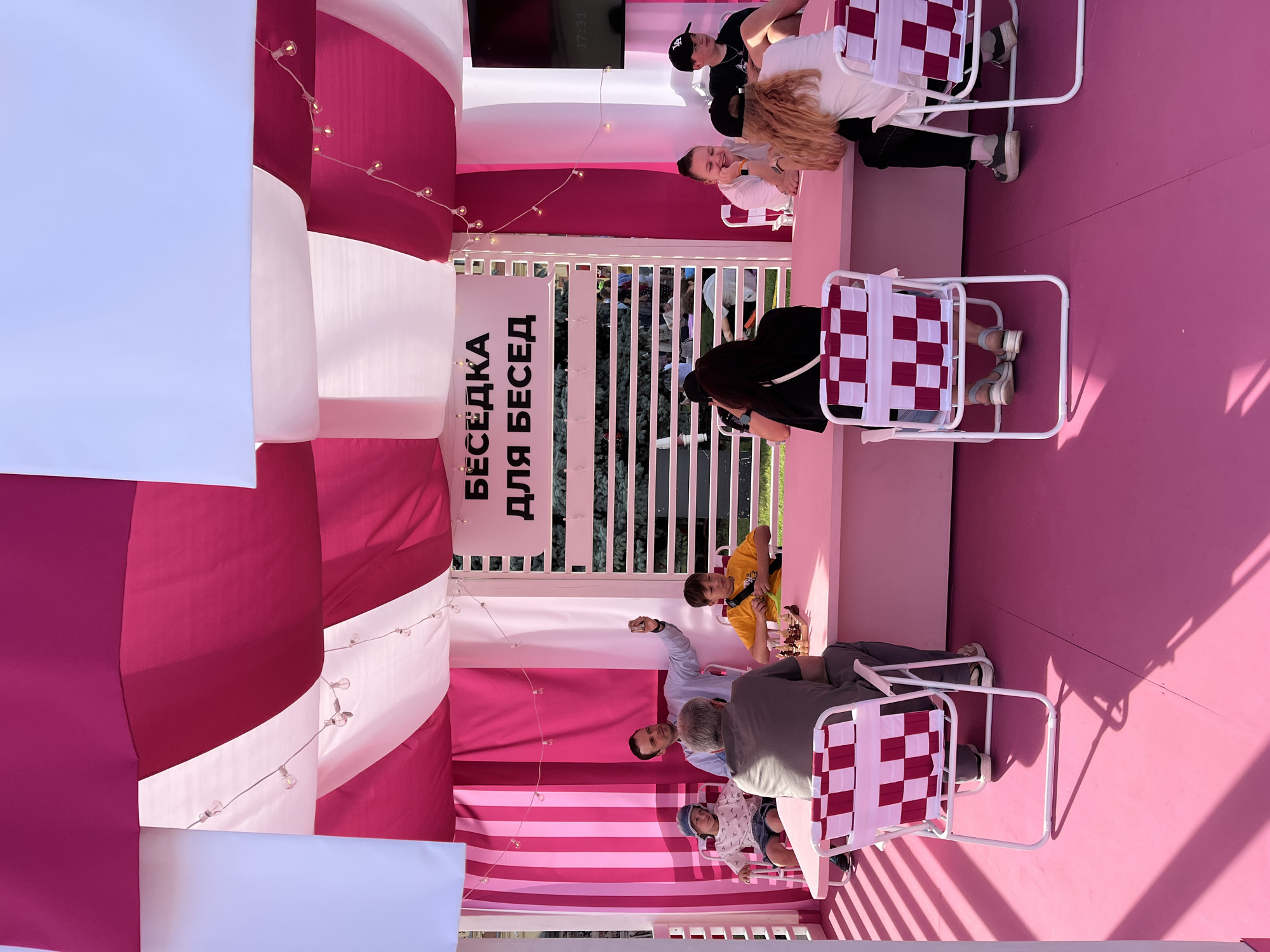 A group of people are gathered under a pink and white striped awning, likely at a public event or park in Nizhny Novgorod, Russia. The awning is decorated with string lights. In the foreground, a pink table is set up, and several individuals are seated around it. One man is holding up a phone, seemingly taking a selfie. A chess game is in progress on the table, with two people actively engaged in it. To the right, a few other individuals are seated and conversing. The floor is also pink. Behind the awning, a white slatted structure with the Cyrillic text "БЕСЕДКА ДЛЯ БЕСЕД" (Gazebo for Conversations) is visible. The overall atmosphere appears to be casual and social.