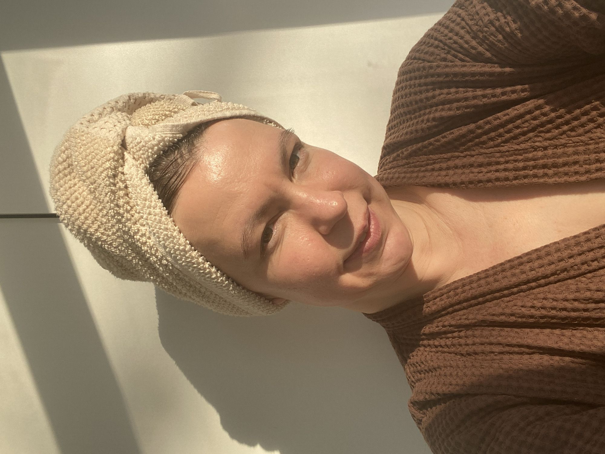 A woman poses for a photograph in what appears to be an indoor setting. She wears a light-colored towel wrapped around her head and a brown waffle-knit robe. The background consists of a plain white wall with a patch of sunlight casting a shadow. The scene appears to be in a private residence. There is no indication that the location is Chaikovsky, Russia.