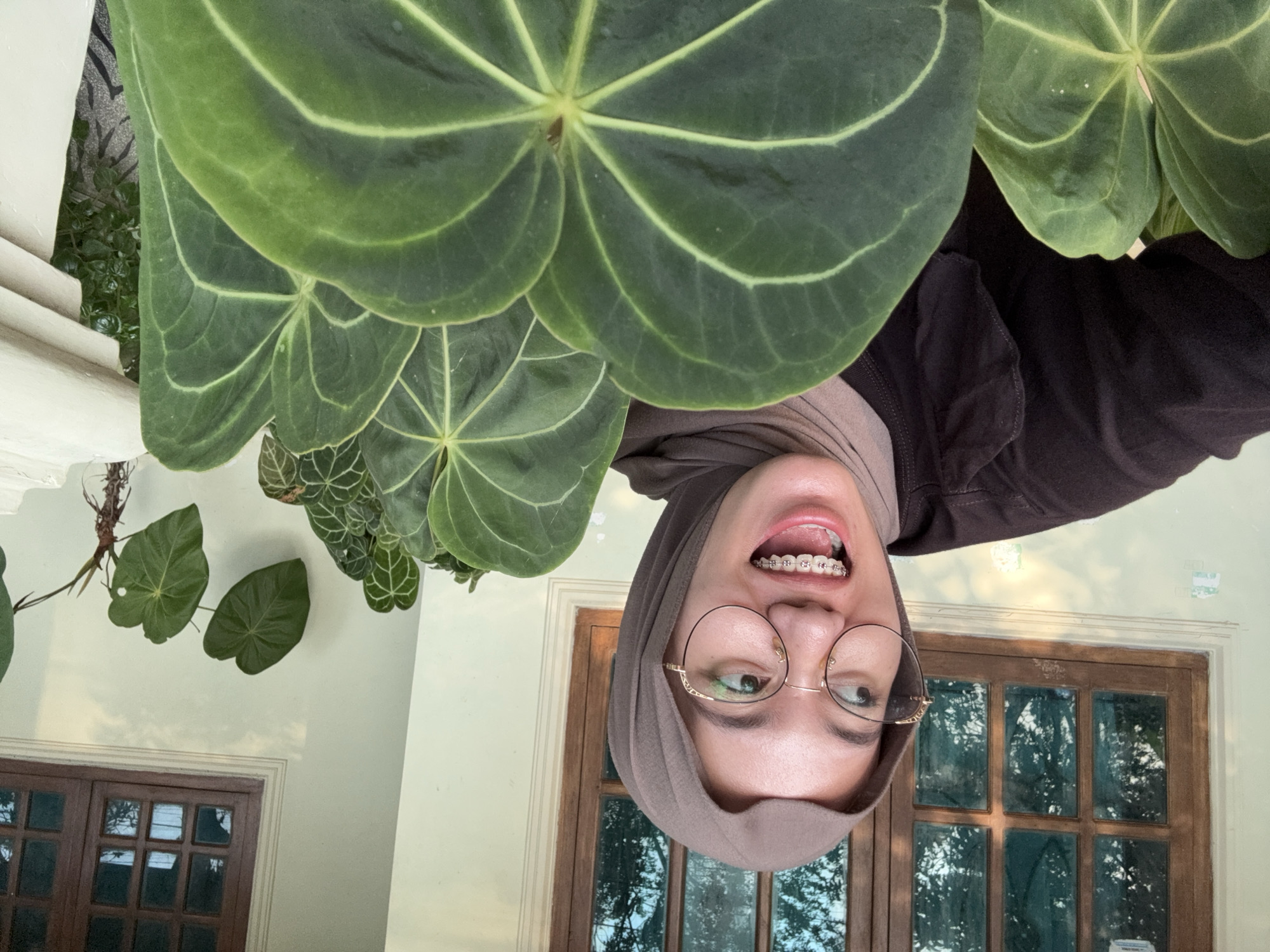 A young woman wearing a hijab and glasses with braces smiles and looks to the left while posing for a photo in Gadingrejo, Indonesia. She is wearing a dark brown top. In front of her are large green leaves. Behind her is a wall with a wooden window.