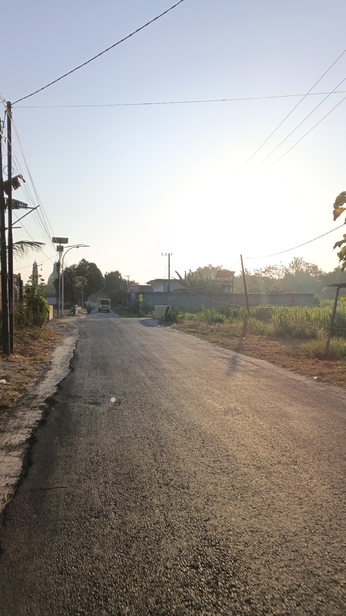 The image shows a road in Soko, Indonesia, on a clear day. The road is asphalt and appears to be in fair condition, with some cracks and unevenness. On the left side of the road, there is a sidewalk with some vegetation and a utility pole. In the distance, there is a small building that looks like a mosque with a dome. On the right side of the road, there is a field with tall grass and some trees. The sun is setting in the background, casting a warm glow over the scene. There is a single-engine vehicle on the road in the distance. The sky is clear with a few thin clouds. The overall impression is one of a quiet, rural setting.