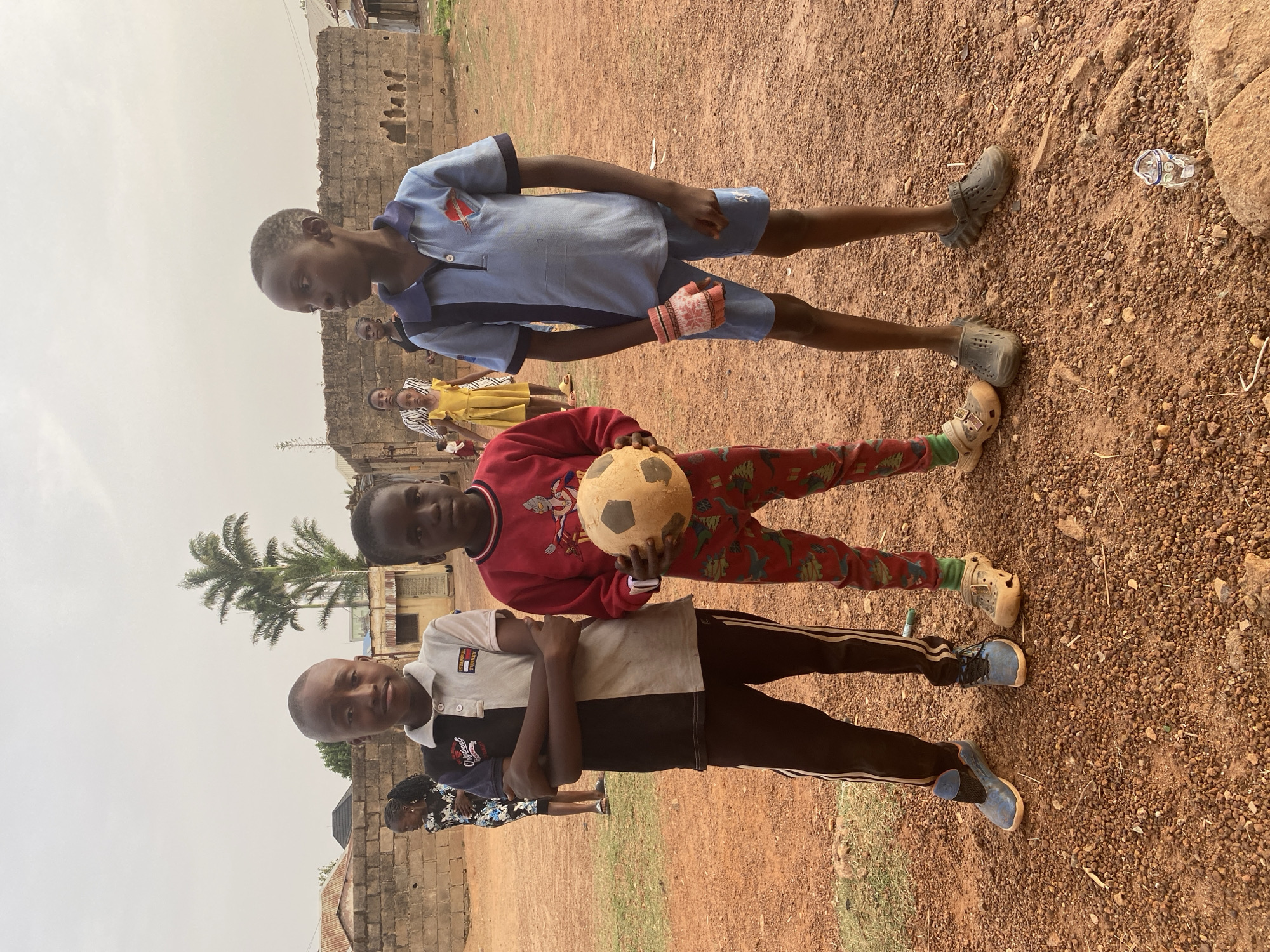 An outdoor scene in Bukuru, Nigeria, captures a group of children gathered on a reddish-brown dirt ground with sparse patches of grass. The sky above is bright but appears overcast or hazy, suggesting daytime. In the immediate foreground, three boys are posing for the camera.
On the left, a boy wears a light grey and black polo shirt with "TOMMY HILFIGER EST. MCMLXXXV" and a small logo visible on the chest. He has black tracksuit pants with white stripes and blue athletic shoes. He offers a slight smile, looking directly ahead.
The central boy holds a well-worn, light brown and black soccer ball in his hands. He is dressed in a red long-sleeve shirt featuring an Ultraman character design, and red and green patterned pants, complemented by light-colored clogs. He looks straight at the viewer with a neutral expression.
To the right, a third boy is dressed in a light blue short-sleeved polo shirt with darker blue accents and a small red and white emblem on the chest, possibly a school uniform. He wears light blue shorts and dark grey clogs, and a single pink and white patterned glove is on his right hand. His gaze is directed downwards and to the left, appearing contemplative or shy.
In the mid-ground and background, other children and adults are partially visible, including a girl in a yellow dress and a woman in a patterned dress. The setting includes simple buildings constructed from concrete blocks or unplastered brick, and several palm trees rise against the muted sky. A crushed clear plastic bottle is visible on the ground to the right. The overall atmosphere suggests a casual gathering, possibly after or before a game of soccer.