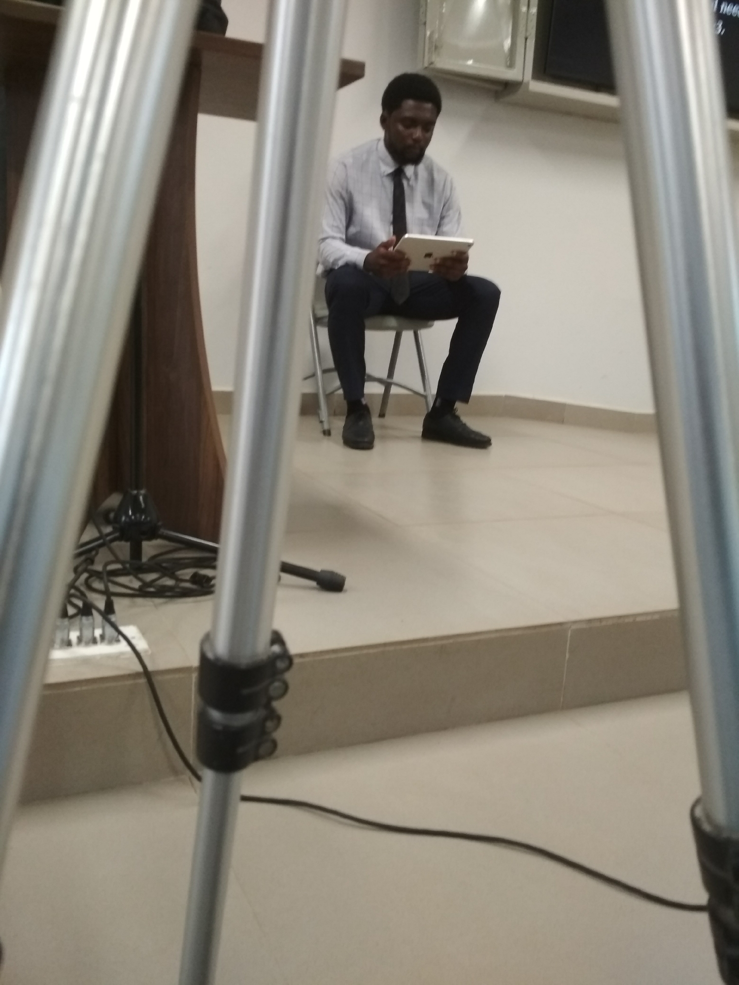 A man of African descent is seated on a chair, holding and looking at a tablet. He is wearing a light-colored, button-down shirt and a dark tie with dark trousers and shoes. He is in what appears to be an indoor space with a podium partially visible to the left. A tripod is prominently positioned in the foreground, partially obstructing the view of the man. There is also a multi-plug power strip and audio cables on the floor to the left. The walls are white. The location is Gura Top, Nigeria.