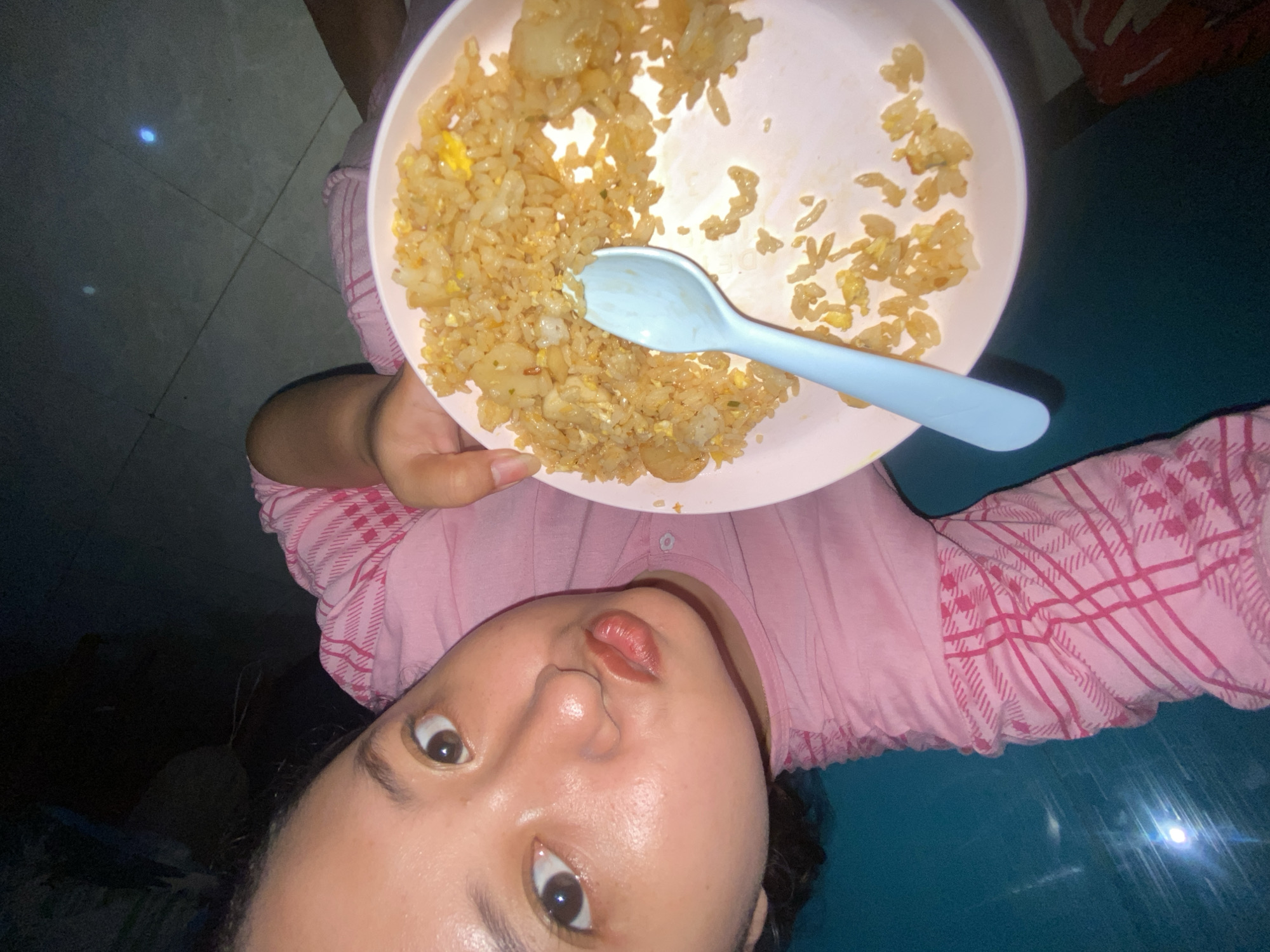 A woman is holding a plate of fried rice and a blue spoon. She is wearing a pink and white checkered shirt. The environment appears to be indoors, with a tiled floor visible in the background. The image does not provide enough information to determine the specific location within Gelam, Indonesia.