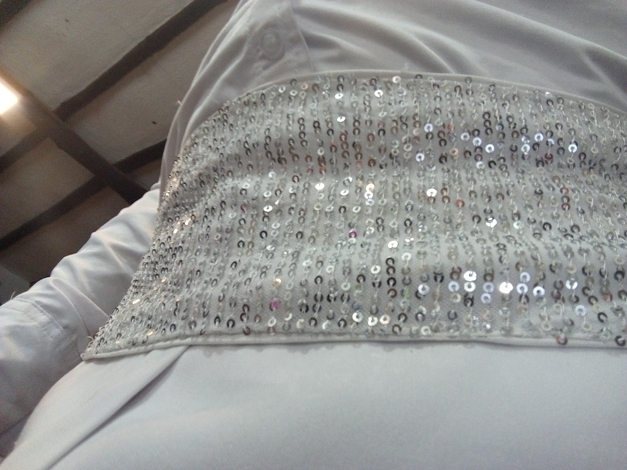 The image shows a close-up view of a person wearing a white garment with a wide, sequined band across the midsection. The sequins are silver and reflect light. The garment appears to be a dress or top. The background is blurred, but there are hints of a ceiling with wooden beams. There is no evidence in the picture to determine the city of Porlamar, Venezuela.