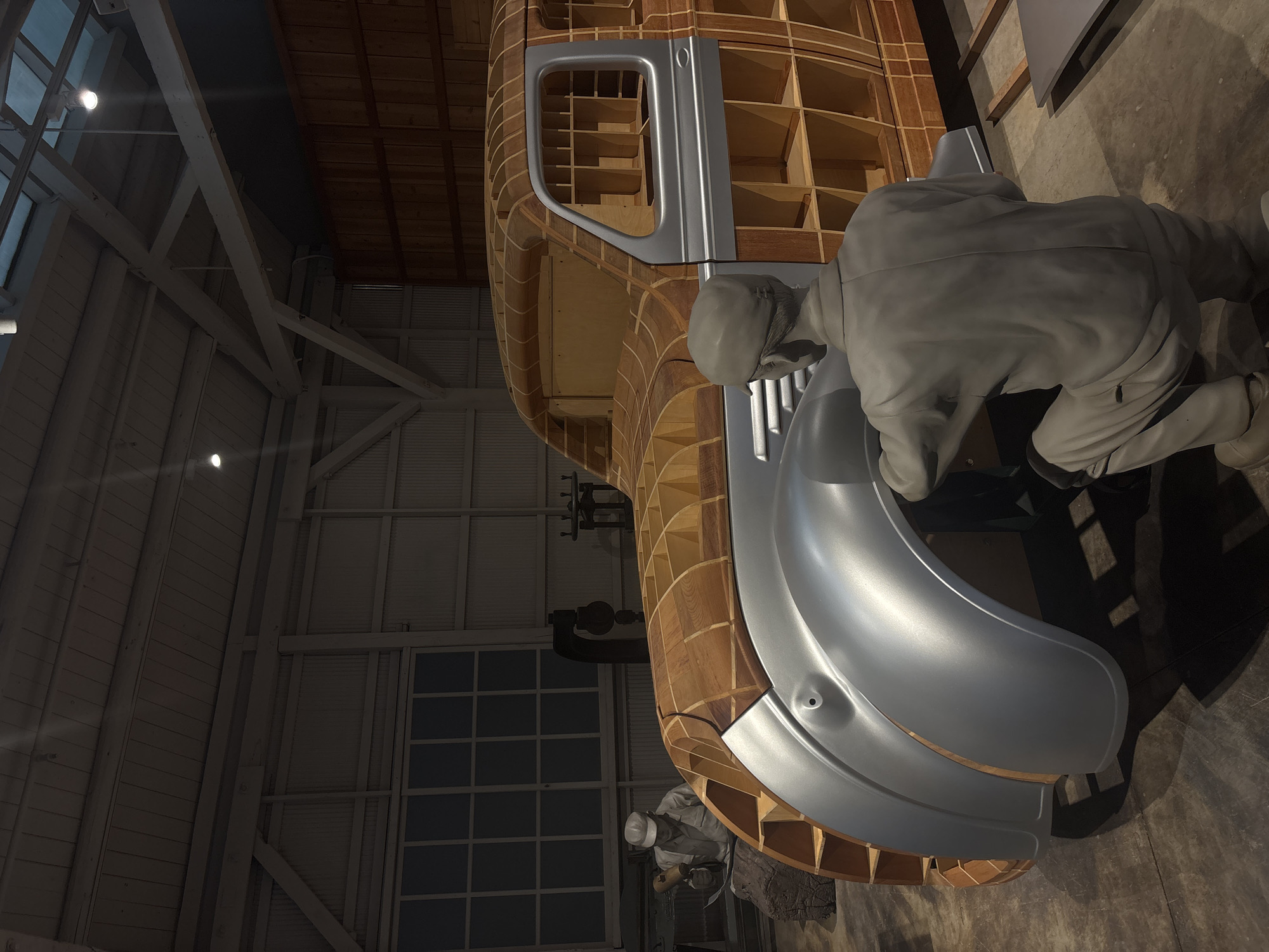 This image depicts a diorama in the Toyota Commemorative Museum of Industry and Technology in Nagoya, Japan, illustrating the automobile manufacturing process. In the foreground, a gray, life-sized mannequin of a man is kneeling and appears to be working on the fender of a car body. The car body itself is under construction, with a wooden frame visible beneath a partially completed silver exterior. Further in the background, another mannequin of a man in a cap is seated and holding a tool, suggesting assembly or shaping work. The environment is an indoor workshop setting, with a concrete floor, corrugated metal walls, and a wooden beamed ceiling with exposed lighting. Large windows with a grid pattern are visible on the back wall, and industrial machinery is present in the mid-ground. The lighting is dim, with spotlights illuminating the main subjects.