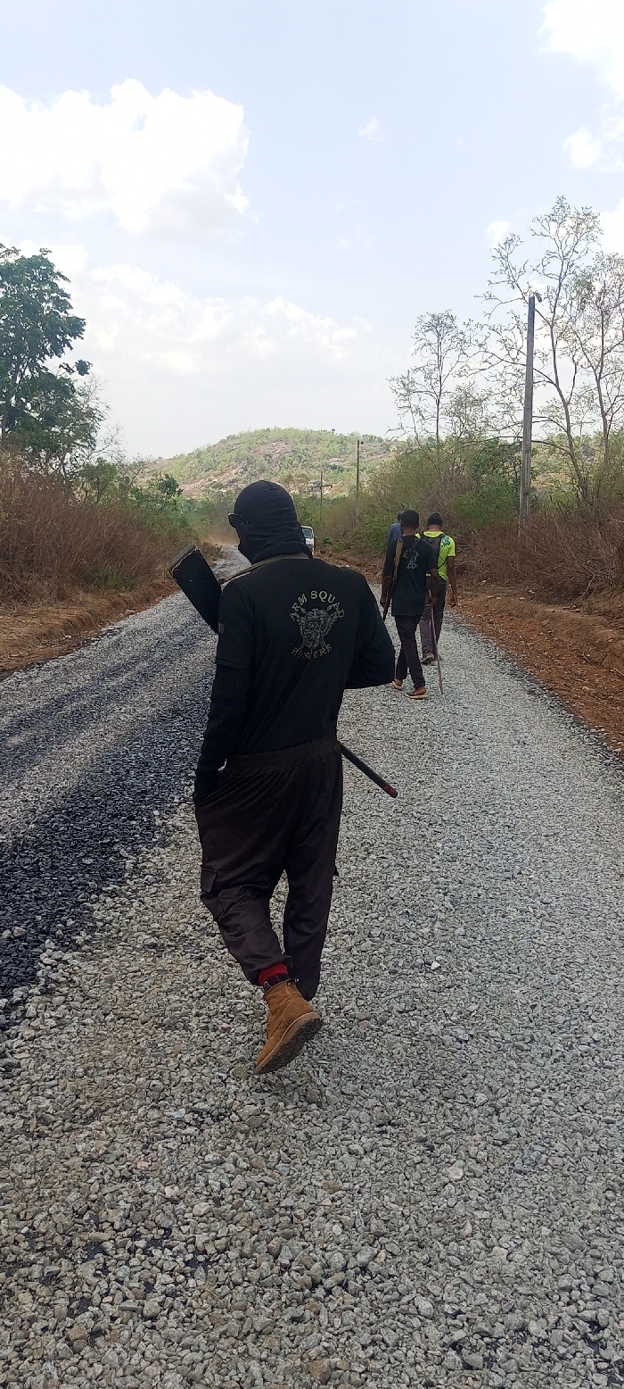 A group of four or more individuals is captured from behind, walking away from the camera on a gravel road under a bright, partly cloudy sky. The setting appears to be rural or semi-rural, characterized by dry, sparse vegetation and a hilly landscape in the background.
The closest individual, central in the frame, wears a black long-sleeved shirt, dark pants (possibly cargo pants), and tan/brown work boots. A red and black checkered pattern is visible on their socks above the boots. Their head is covered by a black balaclava or similar headwear, obscuring their face. On the back of their shirt, a distinct white logo is visible, featuring an emblem that appears to be a winged creature or skull with crossed weapons, above which is the text "ARM SQUAD" and below it, "SLAYERS." This person carries a dark, elongated object resembling a rifle or a long tool over their left shoulder, and another thin, dark object, possibly a sheathed blade or stick, is tucked into their waistband on the right side.
Further down the gravel road, two other individuals are visible. The one directly in front also wears a dark shirt with what appears to be a similar logo on the back, though less clearly discernible. The person further back wears a bright greenish-yellow shirt. All individuals are walking in the same direction.
The road itself is composed primarily of light grey gravel. On the left side of the road, there's a darker, possibly asphalted or freshly laid section of road material, suggesting construction or repair work. In the far distance, a light-colored vehicle, possibly a van or SUV, is faintly visible, stirring up a small dust cloud. The background features several sparse trees and bushes, indicative of a dry environment, with a prominent hill or small mountain range dominating the horizon. A utility pole stands on the right side of the road in the mid-distance. The bright daylight and clear sky suggest it is daytime, likely during a warm period.