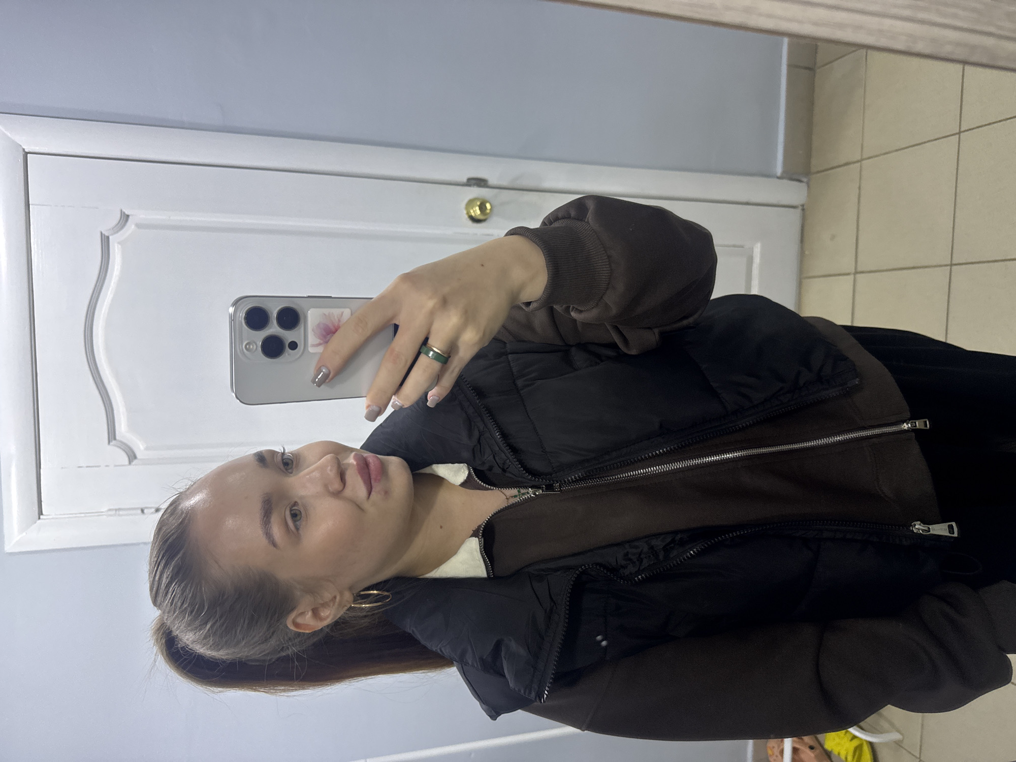 A young woman is taking a selfie in front of a white door. The door is located inside a building with tiled floors. The selfie shows the woman wearing a brown sweatshirt and a black vest. She is holding a smartphone in her left hand, and she has a ring on her left ring finger. The location is Chaikovsky, Russia.