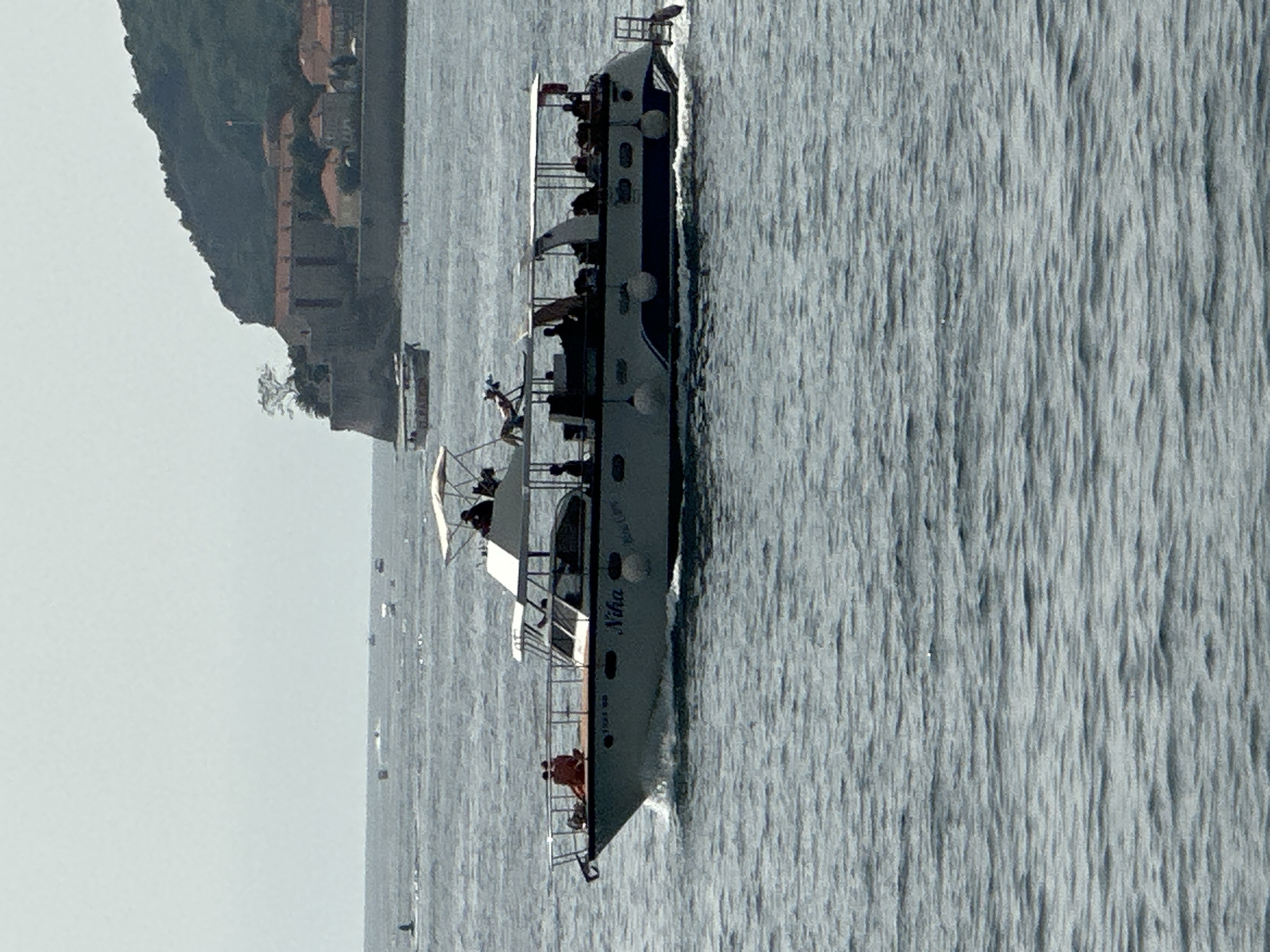 A large tour boat, named "Niha," sails on choppy, grey water, moving from left to right in the frame. The boat has a white canopy over the upper deck, where several passengers are seated. The lower deck is enclosed and has windows. People are visible on both decks, some appearing to be crew members and others passengers. Two individuals sit on the front of the boat, facing away from the camera. The boat has a dark blue stripe along its hull and white fenders. In the background, a rocky, tree-covered hill rises, topped with buildings that appear to be part of an old town, consistent with the location of Budva, Montenegro. The sky is overcast.