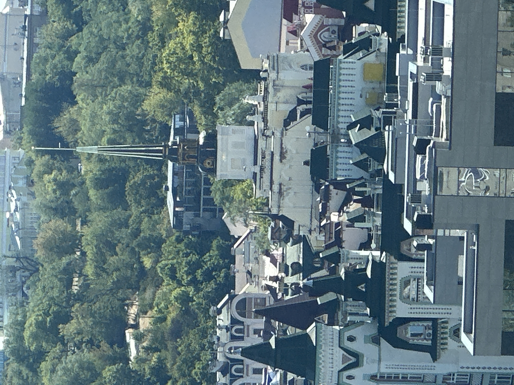This is an aerial, long-shot, eye-level, daytime, outdoor photograph taken in Kyiv, Ukraine, likely from a high vantage point. The image shows a dense urban landscape dominated by a variety of architectural styles. In the foreground, there are several buildings with distinct rooftop features, including pointed, dark green, and grey roofs, some with ornate white trim and decorative elements. One prominent building has a white facade with arched windows and a detailed cornice.
Further back, a white building with a domed structure topped by a tall, slender spire with a flag is visible. This building is situated on a slope surrounded by lush green trees. Behind it, more buildings with varied roofs and facades are visible, some appearing to be older, with classical architectural elements, while others are more modern with flat roofs. The overall impression is of a historic city with a mix of old and new architecture nestled among a verdant, tree-covered hillside. The sky is clear and bright, suggesting a sunny day. No people are visible in the image.