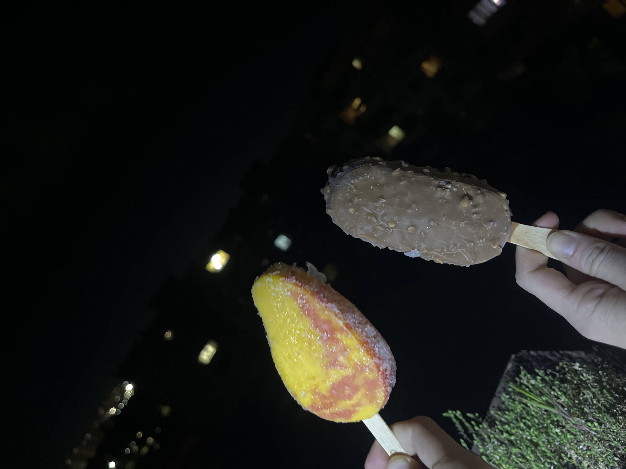 Two ice cream bars on sticks are held in hands. The ice cream bar at the top is chocolate-covered with nuts. The ice cream bar at the bottom is yellow and reddish-orange with a frosted surface. The background is dark and blurred, showing lights of buildings. It is impossible to identify the precise location in Sofia, Bulgaria, without additional information. The scene appears to be at night.