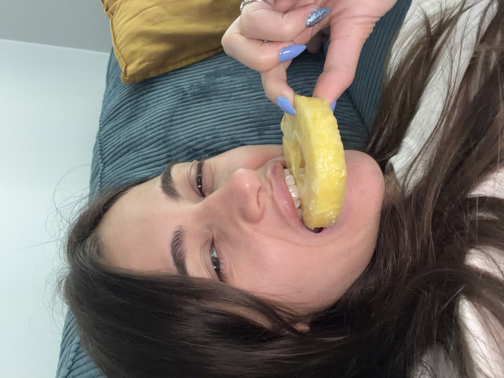 A close-up shot shows a young woman with long, dark hair lying on a couch, about to take a bite of a pineapple ring. Her head is tilted back, and her eyes are open and looking towards the camera. She is holding the pineapple ring with her right hand, which has light blue, stiletto-shaped nails with glitter accents. Her left hand is not visible. The couch appears to be made of corduroy and is a dark teal color, with a mustard-yellow pillow behind her head. The lighting suggests an indoor setting, possibly a living room.