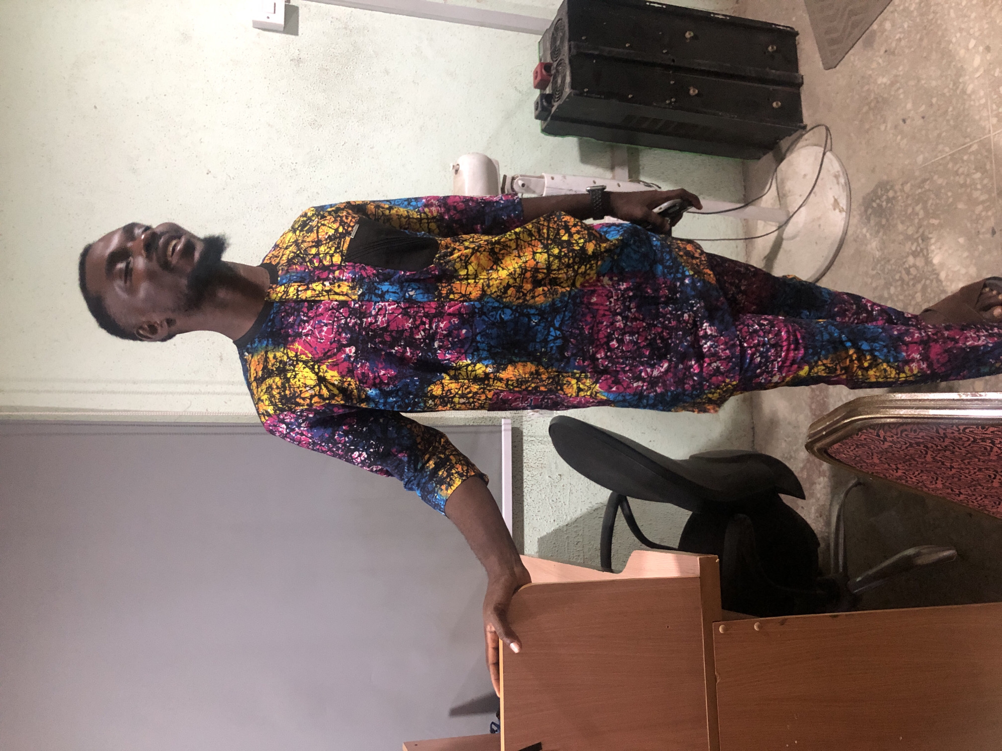 A Black man with a beard and short haircut is standing in what appears to be an office or conference room in Ikeja, Nigeria. He is wearing a colorful dashiki and matching pants, featuring a tie-dye pattern with shades of yellow, blue, pink, and black. His left hand is resting on the edge of a light brown wooden podium. To his right, a black office chair is partially visible. In the background, a white fan with a black cord and a black electronic device are present. The walls are light green and gray, and the floor is speckled concrete. The man is looking to his right and smiling.