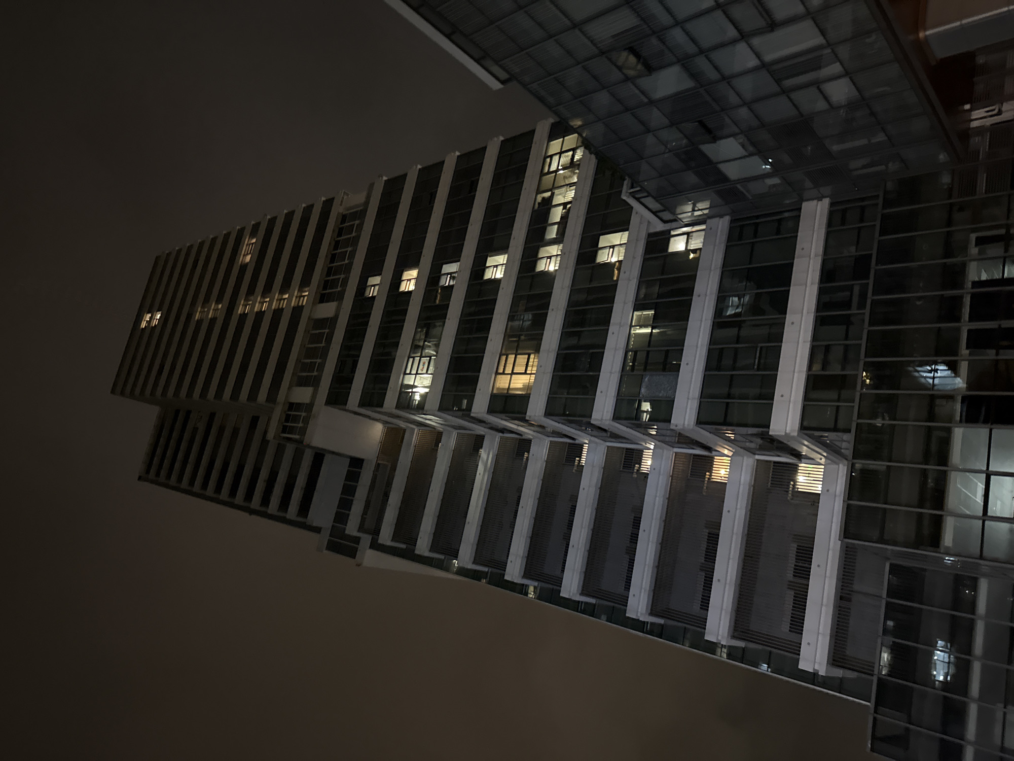 This is a low-angle, full shot of a modern high-rise building at night, located in the Huangpu District of China. The building is tall and slender, with a dark, clouded sky as the backdrop. The facade of the building is a combination of dark glass and white horizontal accents. Many of the windows are illuminated from within, casting a warm glow on the exterior. Some balconies are visible with dark grates. To the right of the main building, a smaller, adjacent structure with a glass facade is partially visible. The overall impression is one of urban nocturnal architecture.
