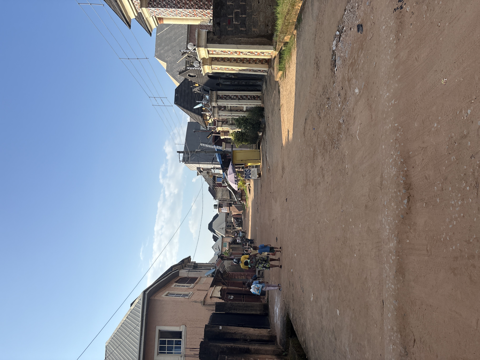 The image depicts a street scene in Obosi, Nigeria, under a clear blue sky. The perspective is from a low angle, looking down a dirt road lined with buildings on both sides. The buildings vary in style and condition, with some appearing to be residential homes and others shops. Many of the structures have decorative elements, and some are adorned with satellite dishes and electrical wires.
Several people are visible in the scene, walking along the road. In the foreground, a group of four individuals are walking together. Further down the street, more people can be seen, some seemingly engaged in daily activities. Laundry is hung out to dry in front of one of the buildings.
The road itself is unpaved, made of reddish-brown dirt, and shows signs of tire tracks and scattered debris. The overall impression is one of a bustling, lived-in community. The lighting suggests it is daytime, likely late morning or early afternoon, given the bright sunlight and clear sky. There is no discernible text visible in the image that provides further context. The scene appears to be a typical day in a Nigerian town.