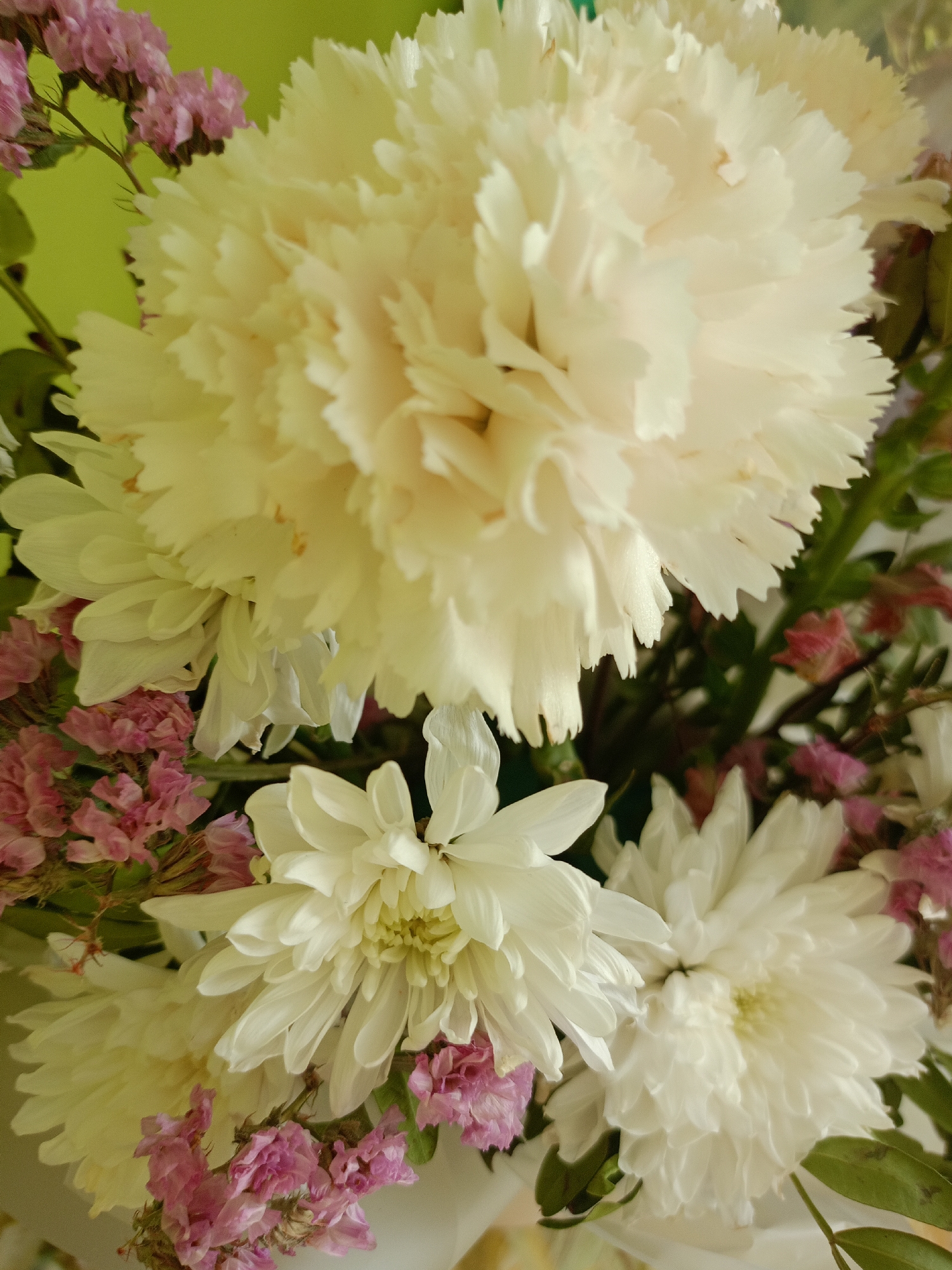 A close-up, overhead shot captures a bouquet of flowers against a light green background. The bouquet features a large, pale yellow carnation prominently in the upper center, its ruffled petals filling the frame. To the lower left and right are two white chrysanthemums with delicate, pointed petals. Small clusters of pink statice flowers are interspersed throughout the bouquet, adding touches of color. The arrangement is densely packed, with some green foliage visible. The lighting is soft, highlighting the textures and layers of the petals. The image is devoid of any human presence or recognizable landmarks, making it impossible to determine its specific location within Moscow, Russia.