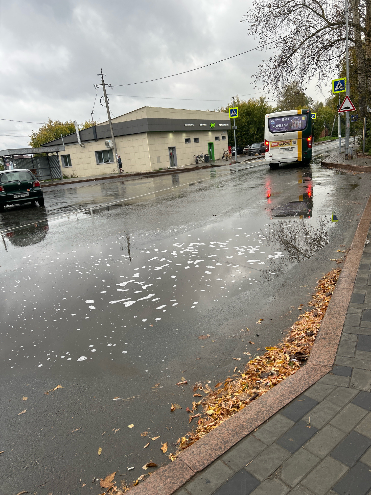 The image depicts a street scene in Tomsk, Russia, likely during an overcast autumn day. The wet asphalt road reflects the cloudy sky and surrounding objects. A green car is parked on the left side of the road, facing away from the viewer. Further down the road, a white and green bus with advertisements on its rear is driving away, its red taillights illuminated. Several pedestrian crossing signs are visible, including one with a yellow diamond shape and a black pedestrian silhouette, and another warning sign with a red triangle and a black zigzag line. A light-colored building with dark gray trim and several windows is situated on the right side of the road. A person is walking along the sidewalk in front of the building. Puddles of water with white foam are visible on the road, and fallen autumn leaves are scattered along the edge of the sidewalk and on the road. The sidewalk is paved with gray and black rectangular tiles. The overall impression is of a damp, cool day with some human activity and traffic on a city street.