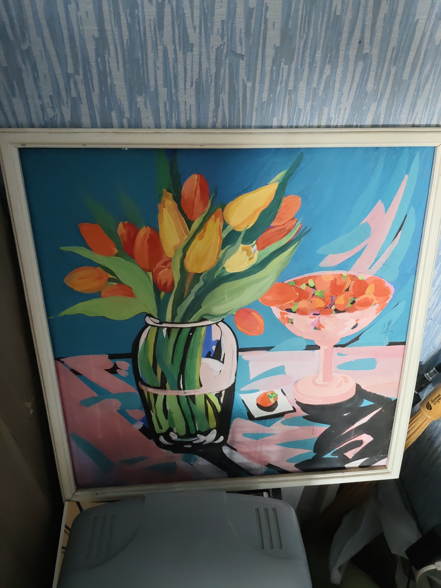The image is a photograph of a framed painting depicting a still life. The painting features a bouquet of orange and yellow tulips in a green-tinted glass vase, placed on a table. To the right of the vase is a pink pedestal bowl filled with orange-colored fruit, possibly apricots or persimmons. A single piece of fruit, similar to the ones in the bowl, sits on a small white square dish in front of the bowl. The background is a vibrant blue with abstract pink and teal shapes. The painting is framed in white and is hung on a wall with light blue textured wallpaper. The bottom portion of the image shows a gray object with vents, possibly a piece of furniture or equipment. The lighting suggests an indoor setting. The image's context as being in Rostov-on-Don, Russia, is not visually evident from the painting or its immediate surroundings in the photograph.