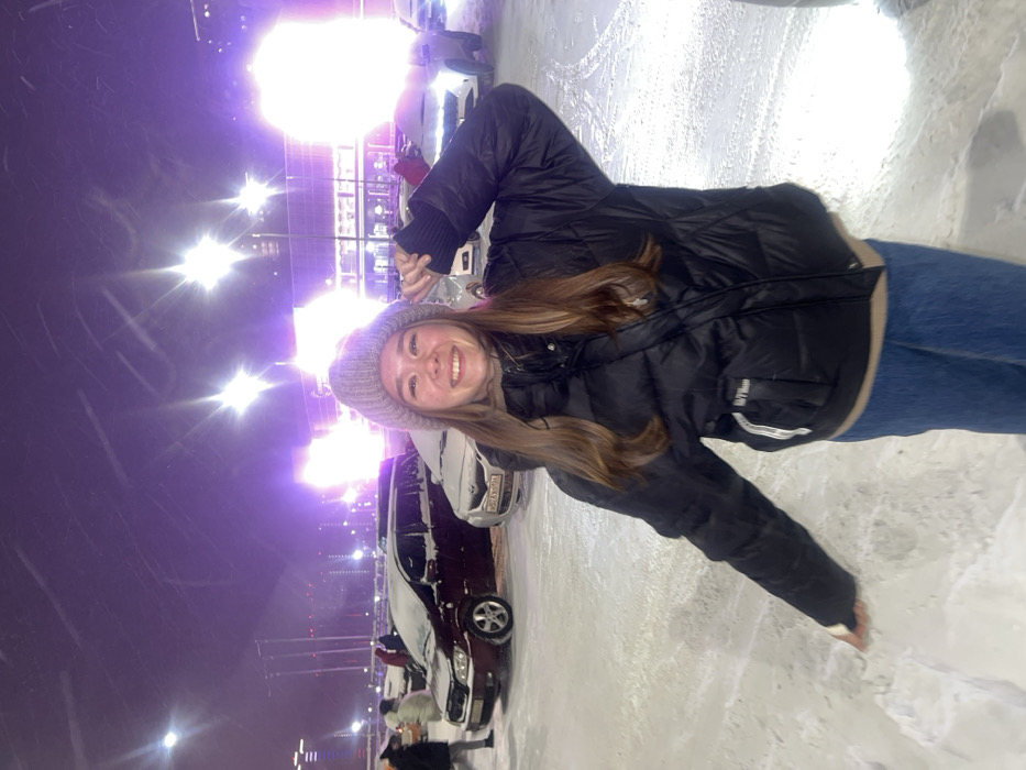 The image displays a young woman smiling broadly in a snowy, outdoor environment at night, illuminated by numerous bright, purple-hued artificial lights. The original image is oriented sideways, with the woman positioned vertically along the right side of the frame.
The central figure is a young woman with long, wavy, light brown hair that falls over her shoulders. She is wearing a light grey knit beanie, a black puffer jacket, and blue jeans. Her jacket appears to have a light beige or cream-colored lining visible around the collar and hem. She is smiling widely, showing her teeth, and looking directly at the camera. Her right hand is raised, with fingers curled, near the side of her head, while her left arm is extended downwards.
The setting is a snow-covered area, likely a parking lot or open plaza, given the presence of parked vehicles and tire tracks visible in the snow. Several cars are parked in the background, partially covered in snow. A prominent dark-colored sedan is visible behind the woman, and a lighter-colored car is parked adjacent to it. The sky is dark, indicating nighttime, and the scene is lit by numerous powerful artificial lights that cast a strong purple glow and create significant lens flares and streaks across the image, particularly in the upper left and right corners. These lights suggest an urban or event setting.
The woman's cheerful expression conveys a sense of happiness and enjoyment. The presence of snow indicates cold, wintery weather conditions.
Visible text includes:
- On the lower right pocket of the woman's black jacket, there is white text that appears to read "O'HARA" or "O'HAVA," accompanied by other smaller, less discernible text.
- On the license plate of the lighter-colored car parked behind the woman, the full plate "016 КР 877 РУС" is clearly visible, indicating a Russian registration.
- A partial license plate on the dark sedan behind her shows numbers ending in "016".