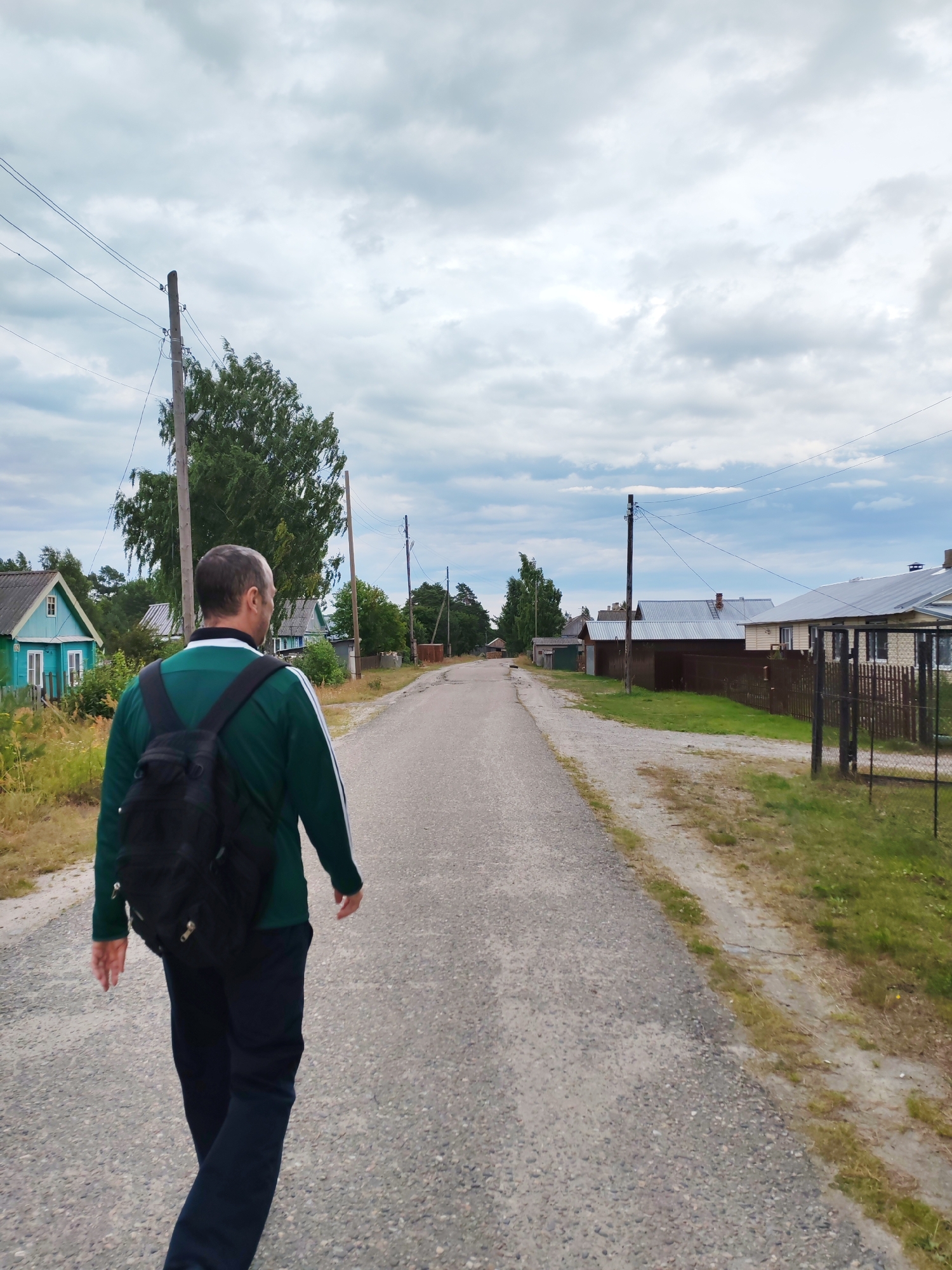 In the city of Shalsky, Russia, a man with a black backpack walks away from the camera on an asphalt road. He is wearing a green jacket with white stripes on the sleeves and dark pants. The road is lined with houses and trees on both sides. The sky is cloudy. The scene appears to be a rural or semi-rural setting.