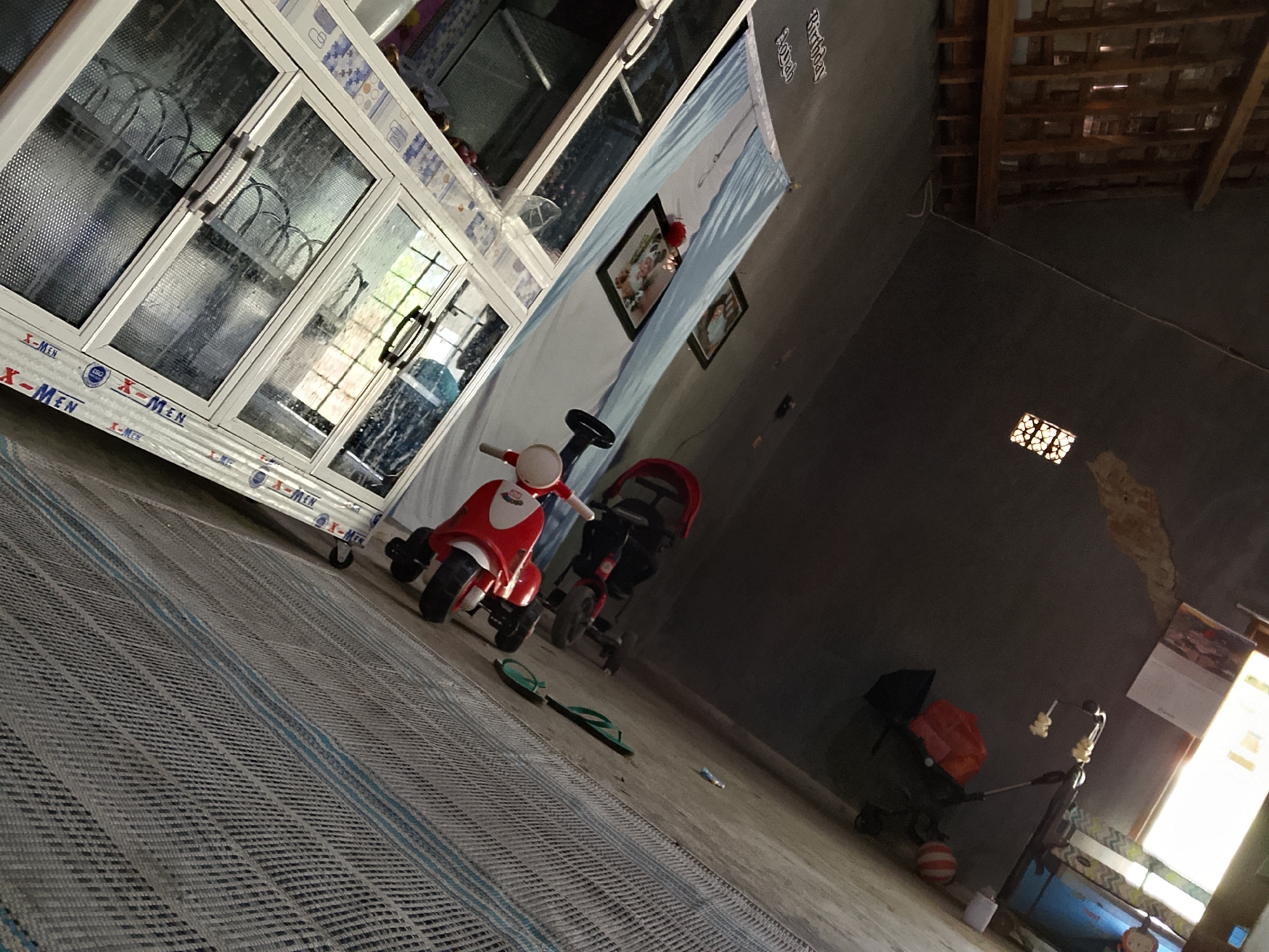 In a dimly lit room in Besuki, Indonesia, a white cabinet with glass doors stands against a wall adorned with a large blue and white banner featuring a landscape. On the floor in front of the cabinet are two children's toys: a red and white tricycle and a red stroller. A pair of green flip-flops lies on the floor near the tricycle. The room has a concrete floor and a wooden beamed ceiling. To the right, a window lets in bright sunlight. Further back, a different stroller with an orange bag is visible, along with a small orange and white striped ball. A white tube-like object is also present on the floor. The overall scene suggests a domestic setting, possibly a storage area or a children's play space, with a slightly cluttered appearance.